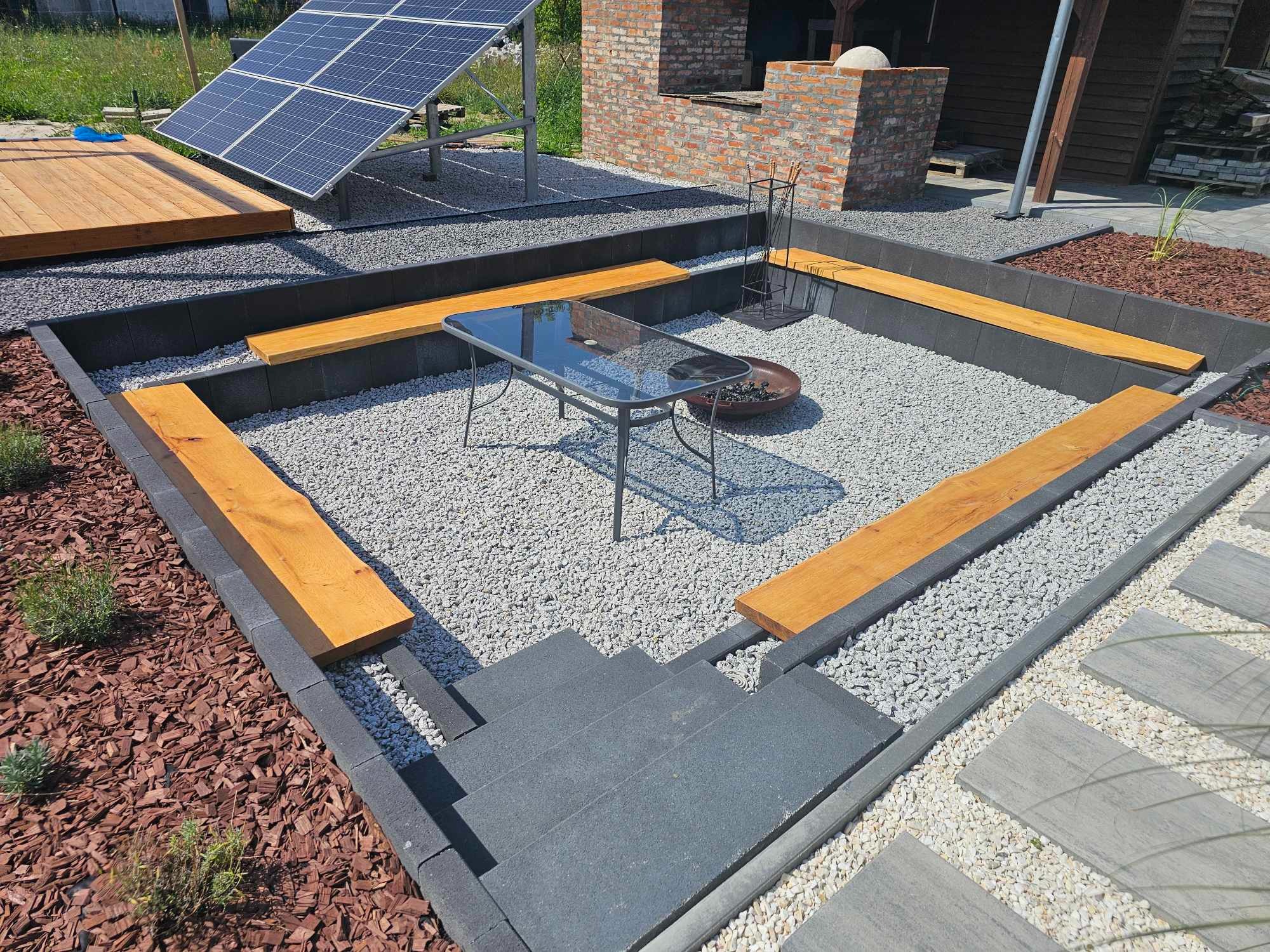 Nowoczesna aranżacja przestrzeni zewnętrznej z paleniskiem otoczonym szarym kamieniem, drewnianymi siedziskami i szarą kostką, w tle panel słoneczny i murowany grill.