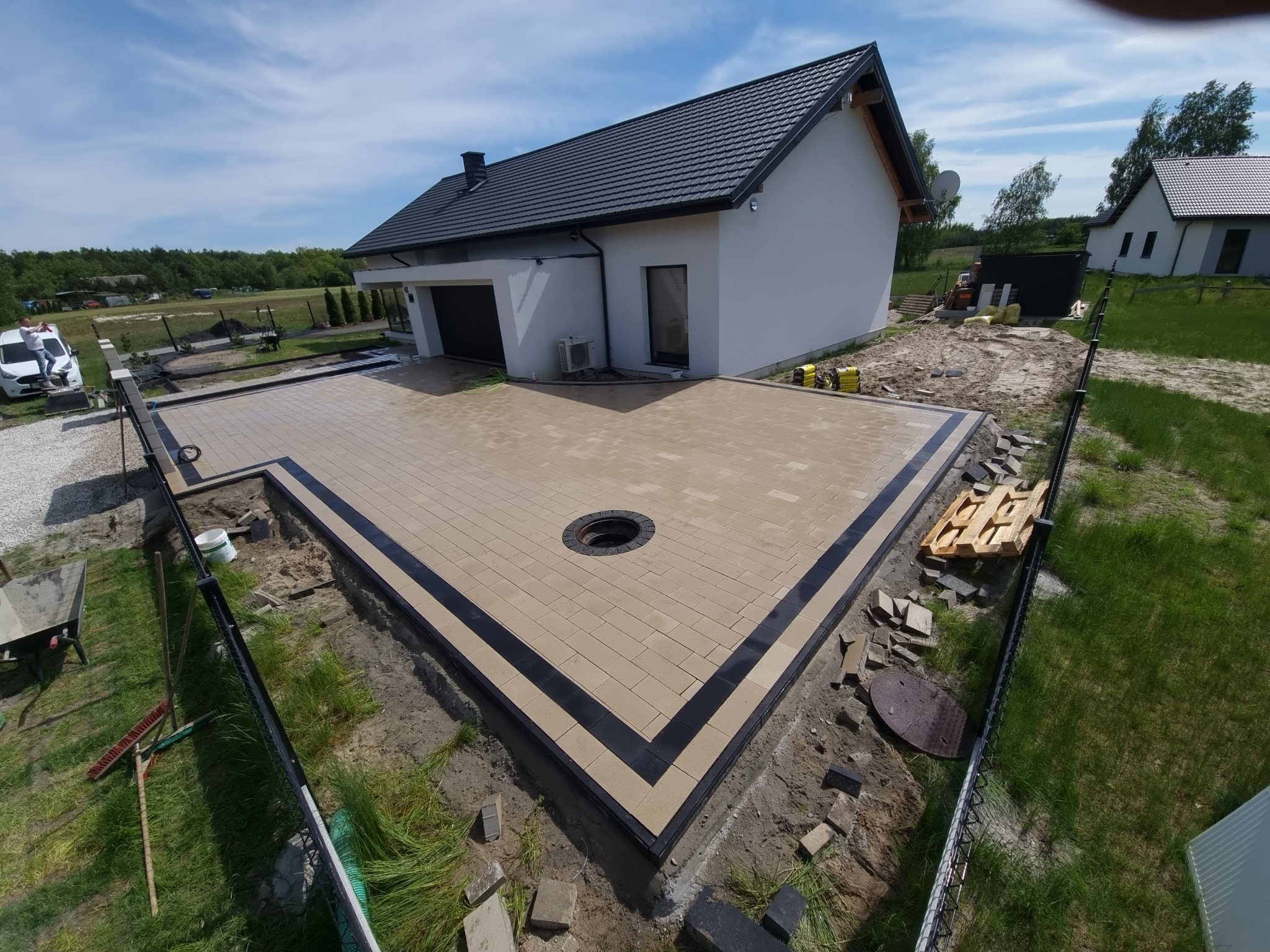 Nowo ułożona, beżowo-czarna kostka brukowa na podjeździe przed nowoczesnym domem z czarnym dachem, widok z góry na studzienkę kanalizacyjną wkomponowaną w kostkę.