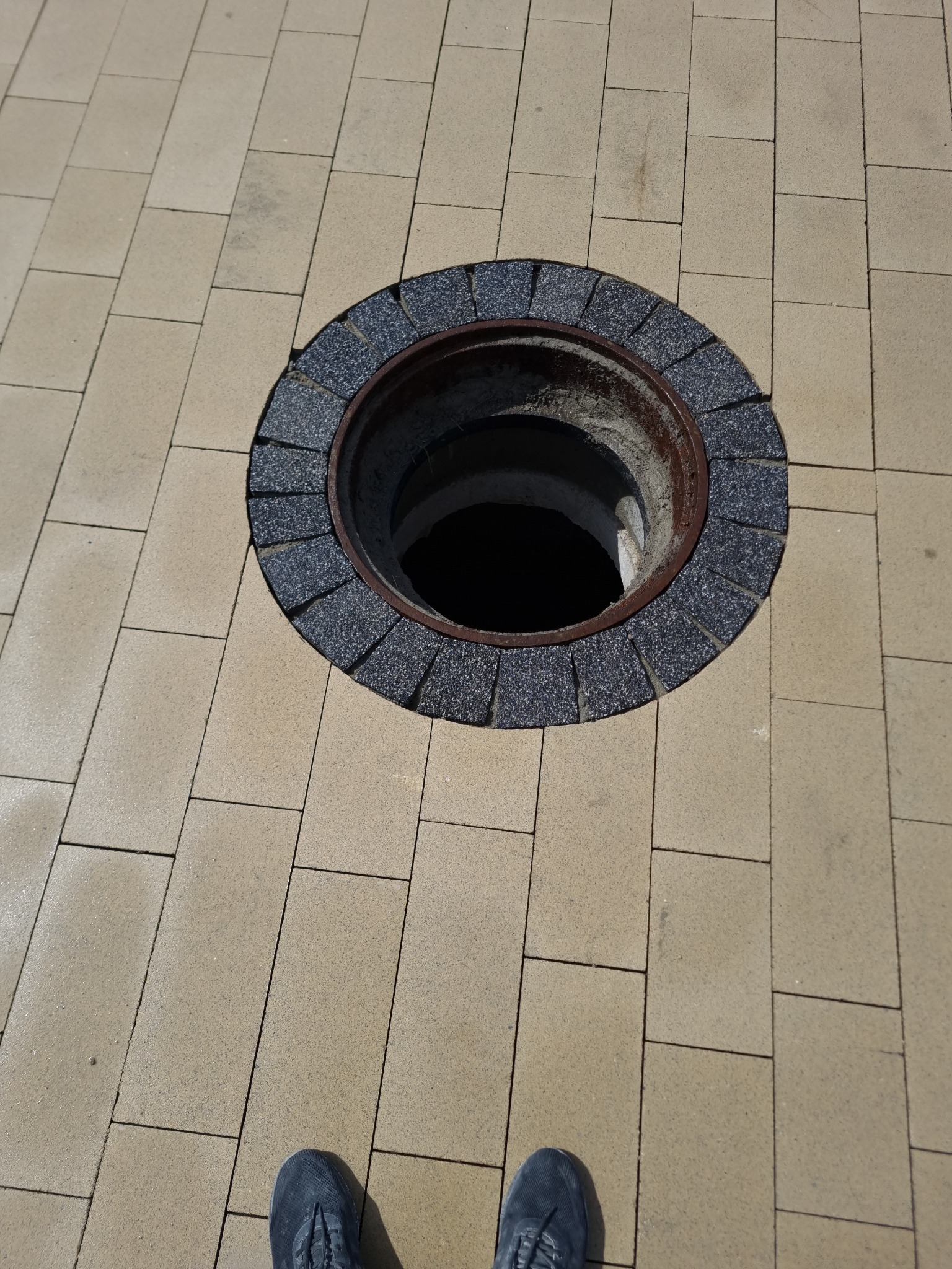 Otwarty właz kanalizacyjny otoczony ciemnoszarą kostką granitową, wkomponowany w jasną, prostokątną kostkę brukową. W dolnej części kadru widoczne stopy osoby fotografującej w szarych butach...