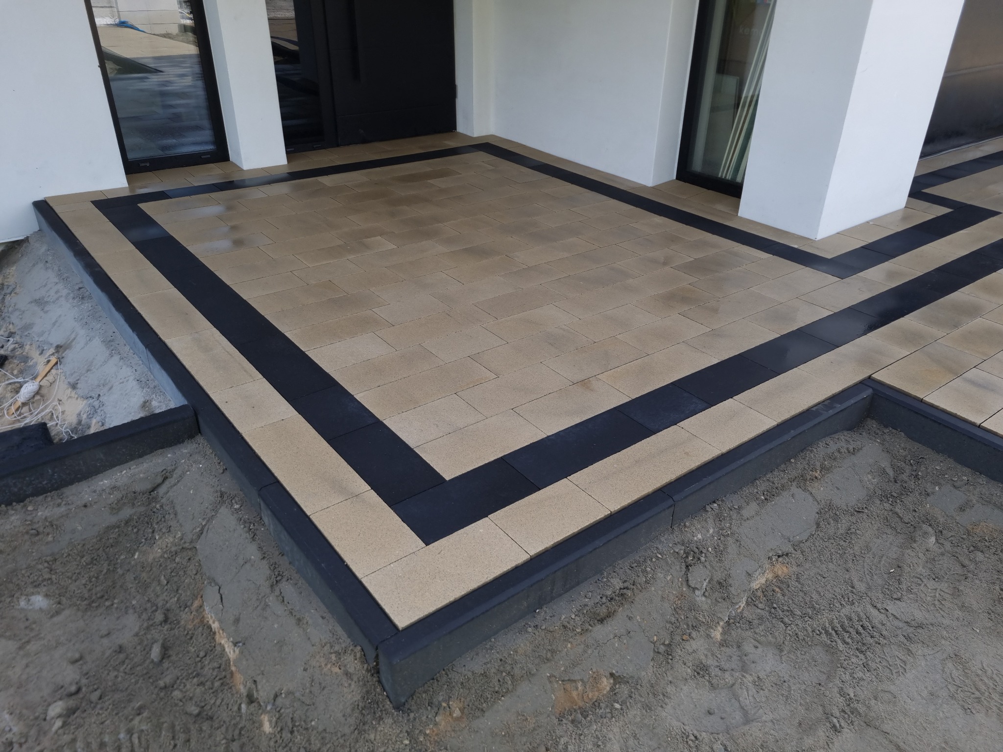 Wykończony taras z jasnej kostki brukowej z czarną obwódką, widoczne niedokończone prace przy krawężniku i przygotowane podłoże.