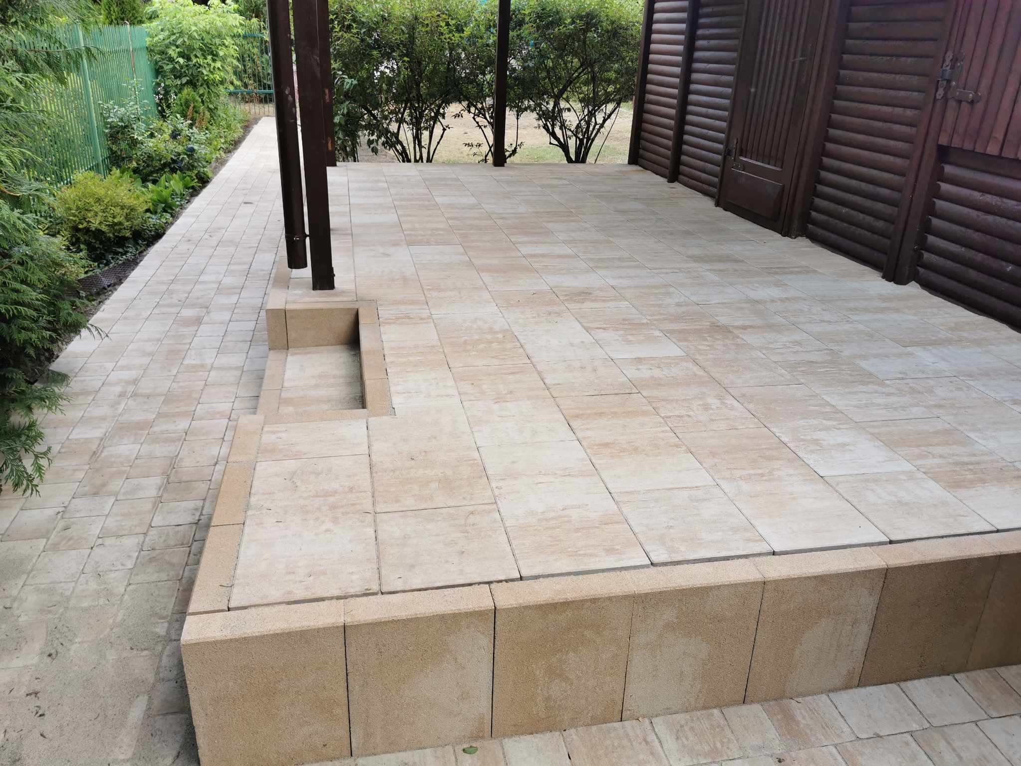 Beżowa nawierzchnia z kostki brukowej tworząca taras przy drewnianym budynku, z widocznymi elementami zieleni w tle.
