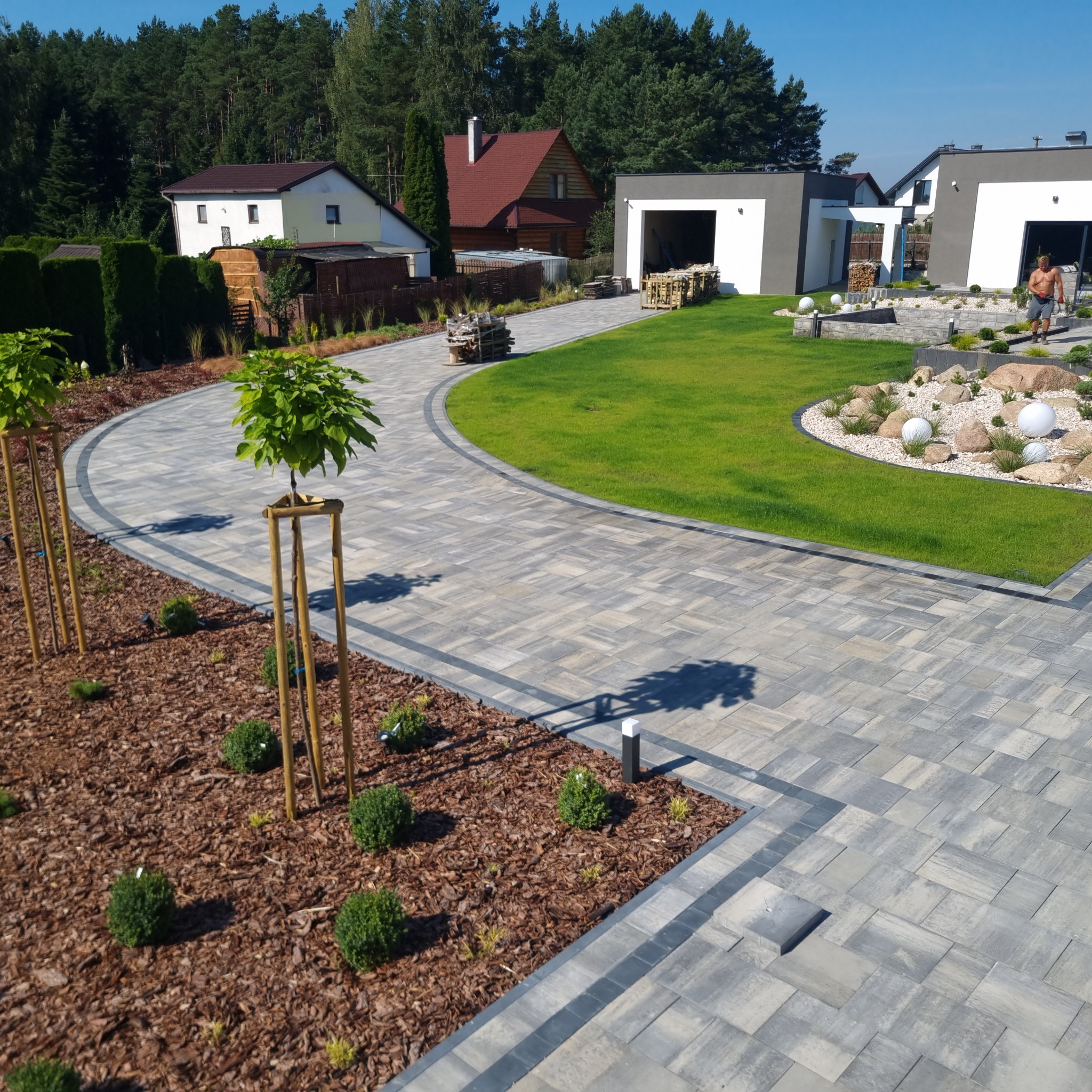 Nowoczesny podjazd z szarej kostki brukowej ułożonej w łuk, obrzeża z ciemniejszej kostki, rabaty wysypane korą z młodymi drzewkami i krzewami, trawnik z dekoracyjnymi kamieniami i kulami, w tle...