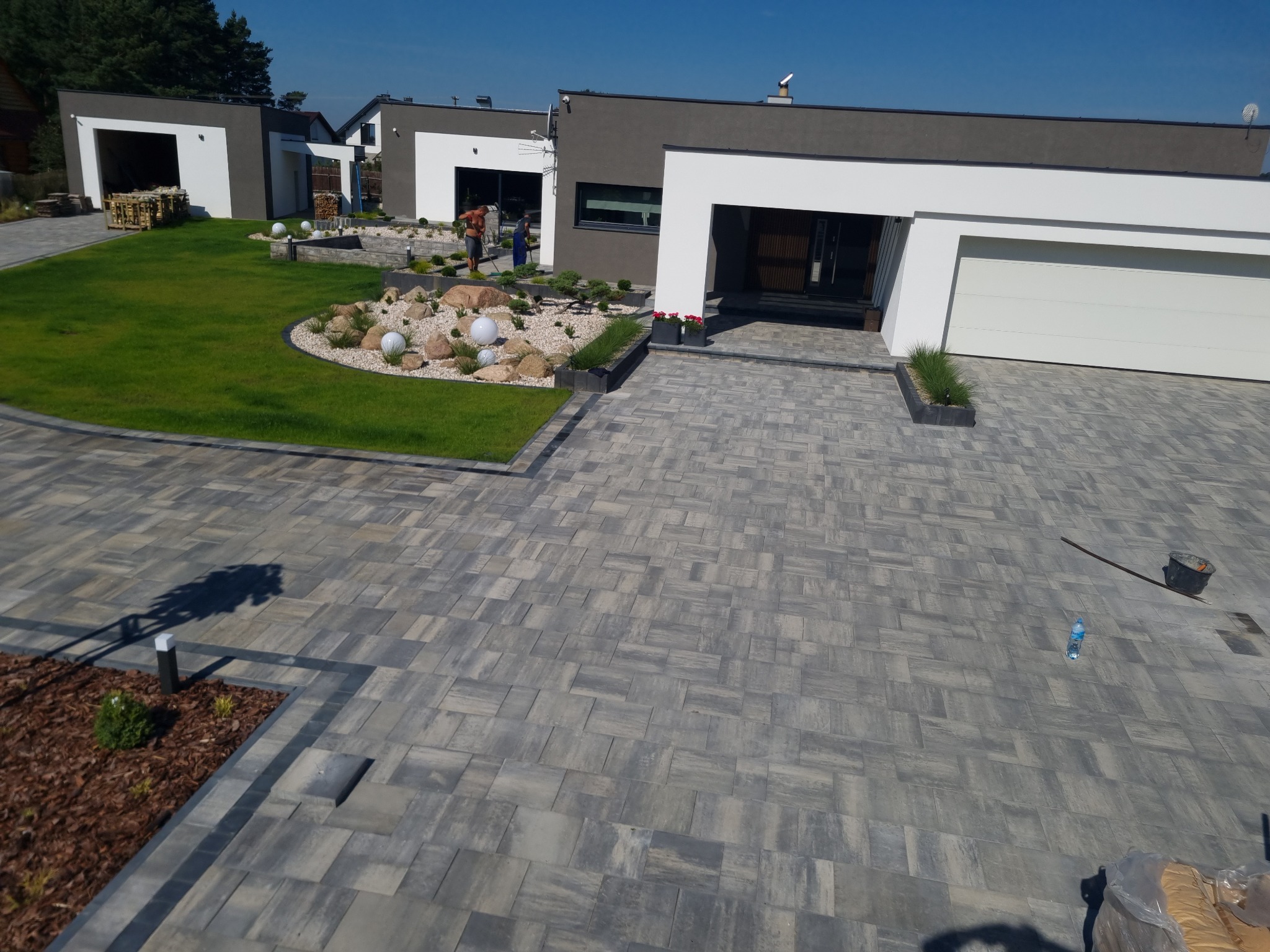 Szeroki kadr na nowoczesny dom z szarej kostki brukowej na podjeździe, z elementami architektury ogrodowej, takimi jak trawnik z białymi kulami i rabaty z kamieniami, oraz dwoma pracownikami w tle.