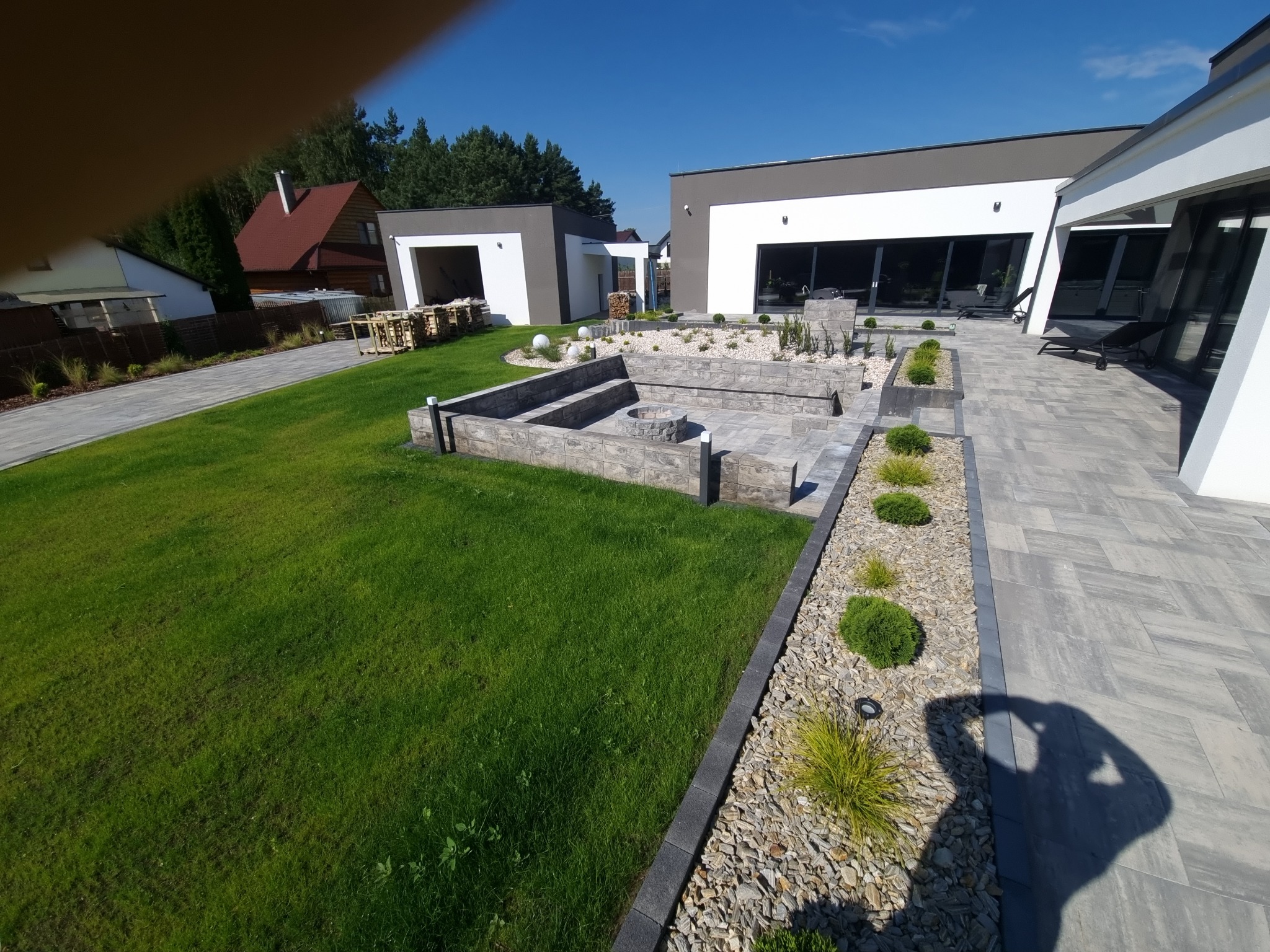 Nowoczesny ogród z wybrukowanym tarasem i ścieżkami, elementami dekoracyjnymi z kamienia i zieleni. Widoczna strefa relaksu z paleniskiem obudowanym kamienną konstrukcją, trawnik i budynki w tle.