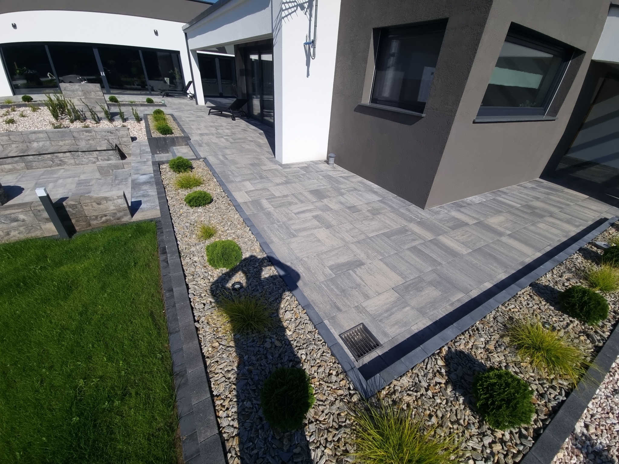 Ułożona kostka brukowa w odcieniach szarości przed nowoczesnym domem z dużymi oknami, obrzeża z kamieni ozdobnych i niskich roślin, cień fotografa widoczny na kostce.