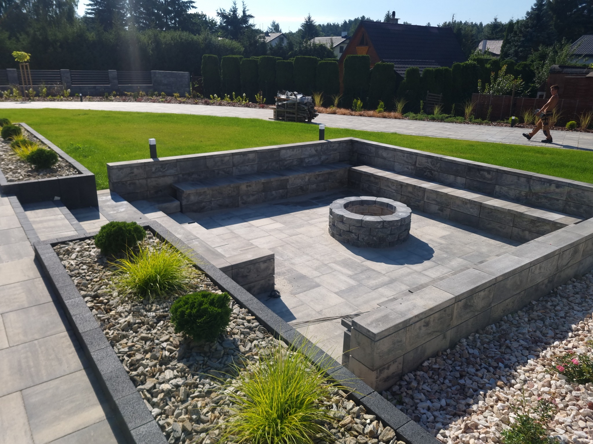 Ogród z paleniskiem otoczonym kamienną konstrukcją z siedziskami, wyłożony kostką brukową w odcieniach szarości, z fragmentami rabat wysypanych kamieniami i obsadzonych roślinami ozdobnymi, na tle...