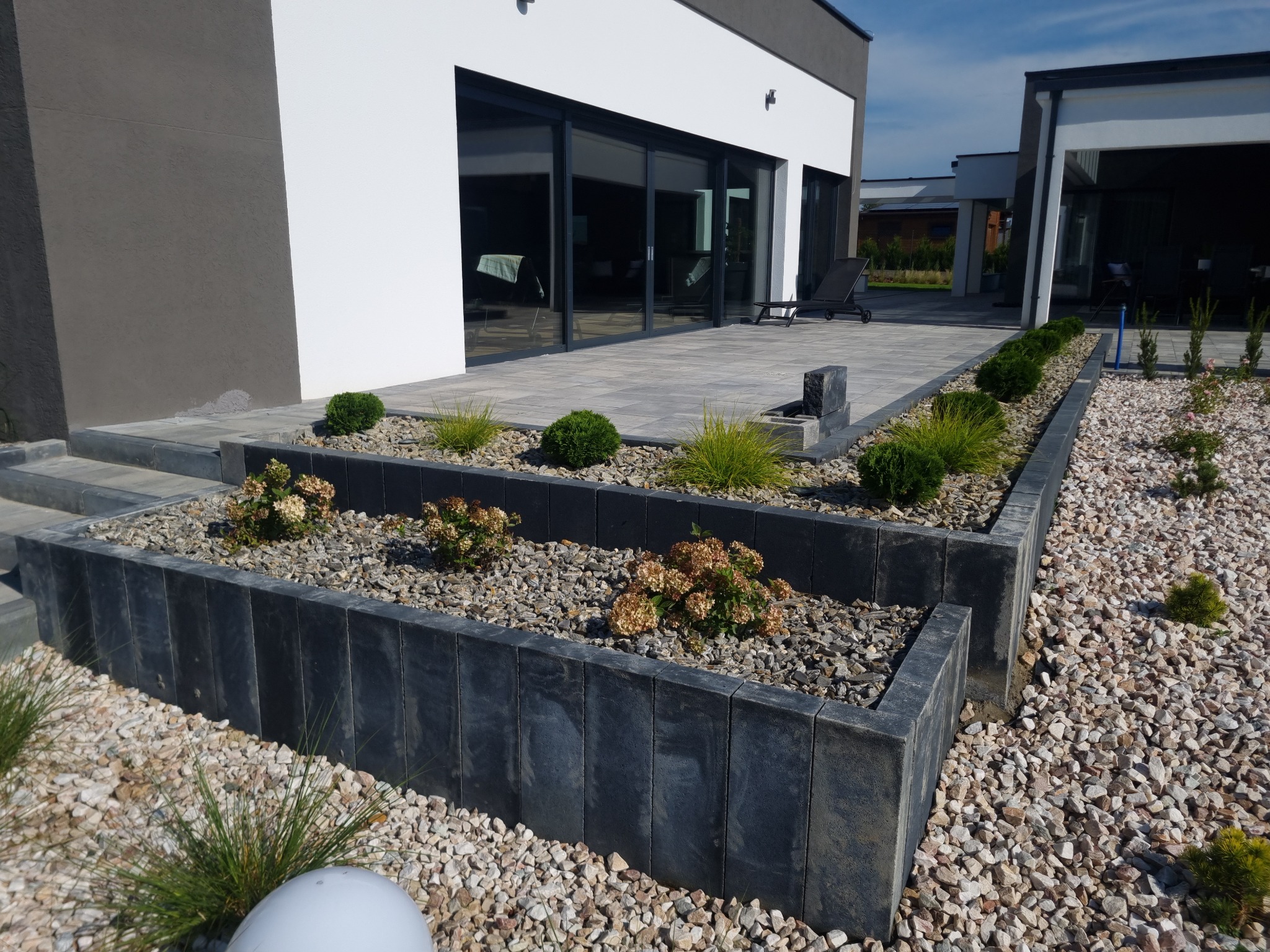 Nowoczesny taras z szarej kostki brukowej, obrzeża z ciemnych betonowych elementów, rabaty z kamykami i roślinami, widok na przeszklone drzwi przesuwne i minimalistyczny budynek w tle.