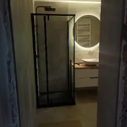 Nowoczesna łazienka z czarną kabiną prysznicową typu walk-in, umywalką nablatową na drewnianym blacie i okrągłym lustrem z podświetleniem LED, widoczna z perspektywy wejścia do pomieszczenia.