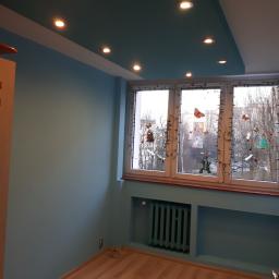 Cegiełka Krzysztof Pachniewicz - Pomalowane na niebiesko wnętrze pokoju z drewnianą podłogą, oświetleniem punktowym w suficie i oknem ozdobionym naklejkami i lampkami.