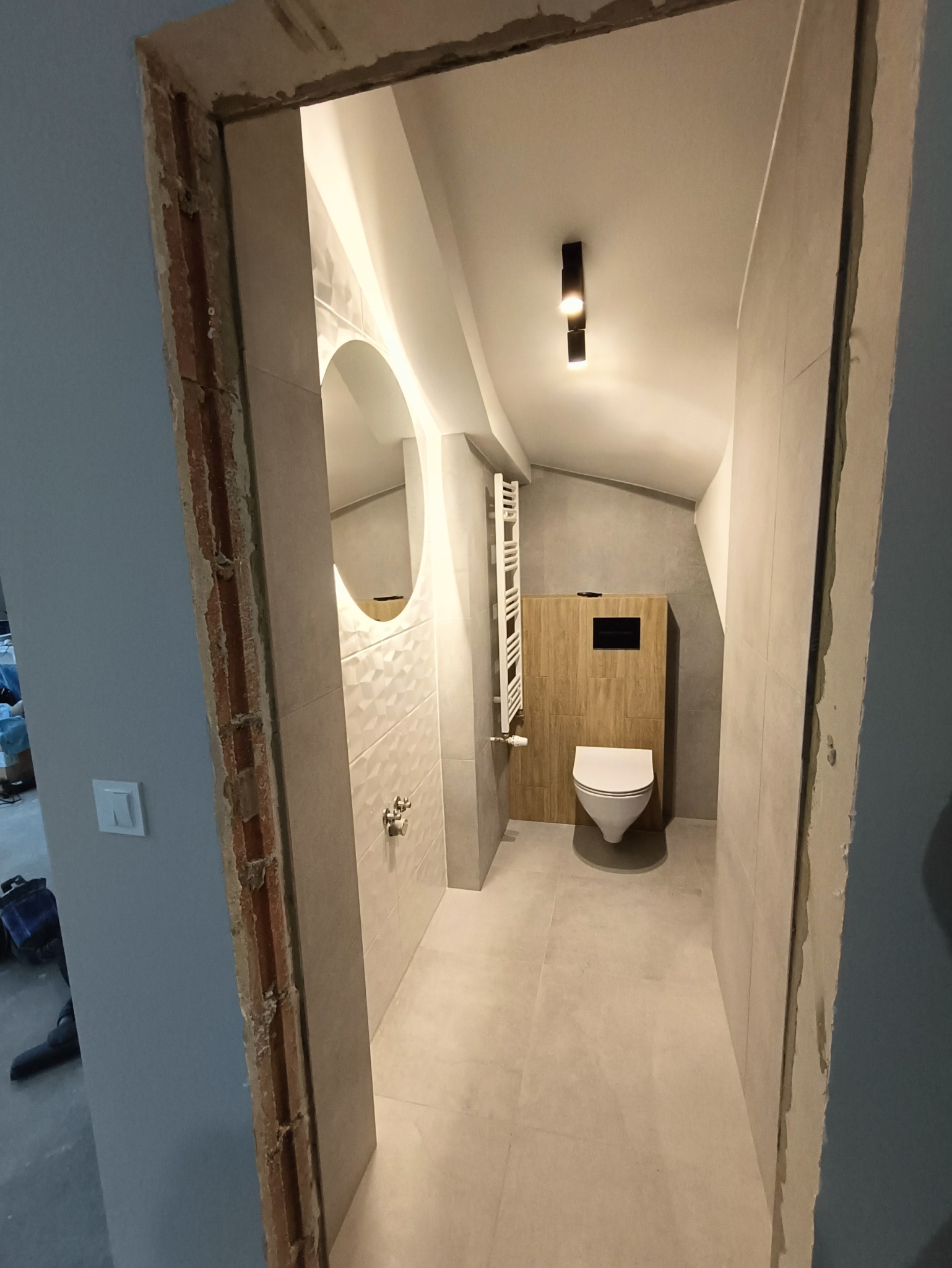 Nowoczesna łazienka na poddaszu z umywalką z okrągłym lustrem, grzejnikiem drabinkowym i podwieszaną toaletą na tle drewnianej zabudowy, widok z korytarza z surowym wykończeniem ościeżnicy.