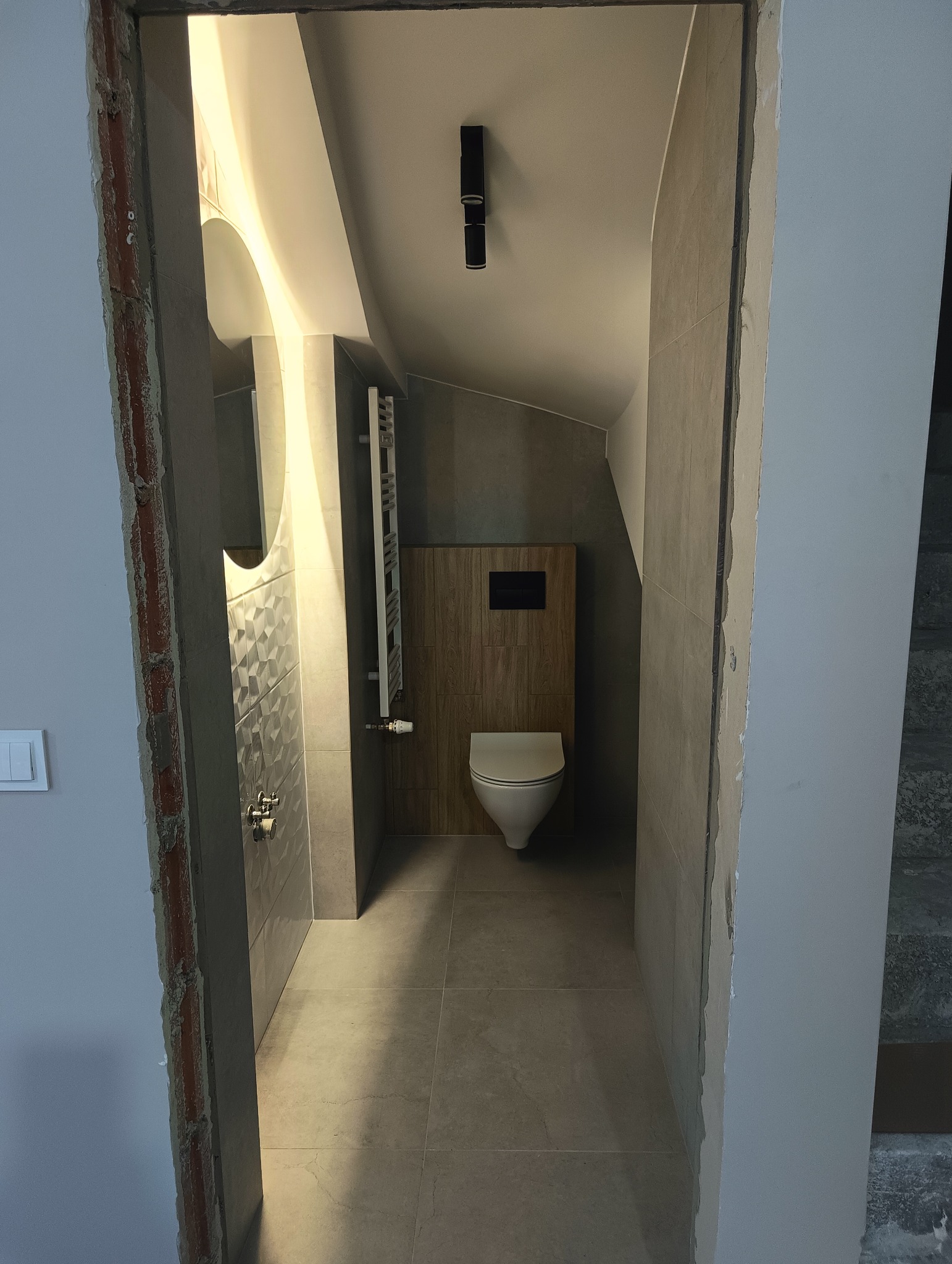 Wnętrze łazienki na poddaszu z toaletą typu Geberit, grzejnikiem drabinkowym i owalnym lustrem z podświetleniem LED, widziane z korytarza przez surową framugę drzwi.