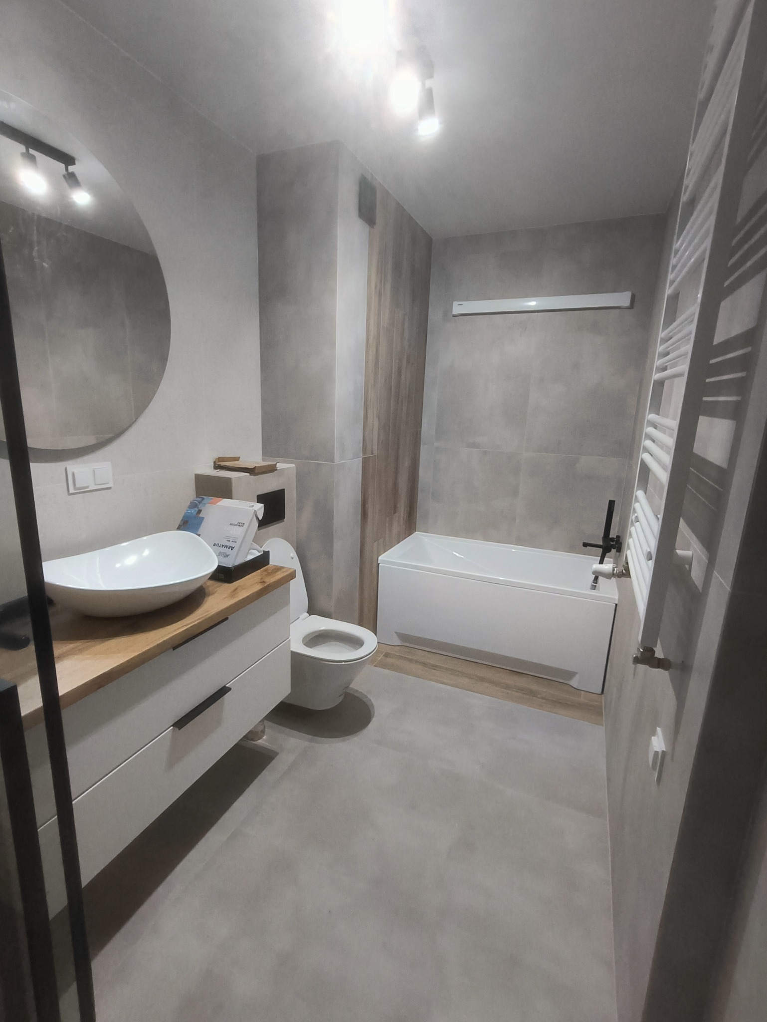 Nowoczesna łazienka w szarościach z drewnianymi akcentami, owalna umywalka nablatowa, biała wanna i drabinkowy grzejnik, widoczne opakowania armatury.