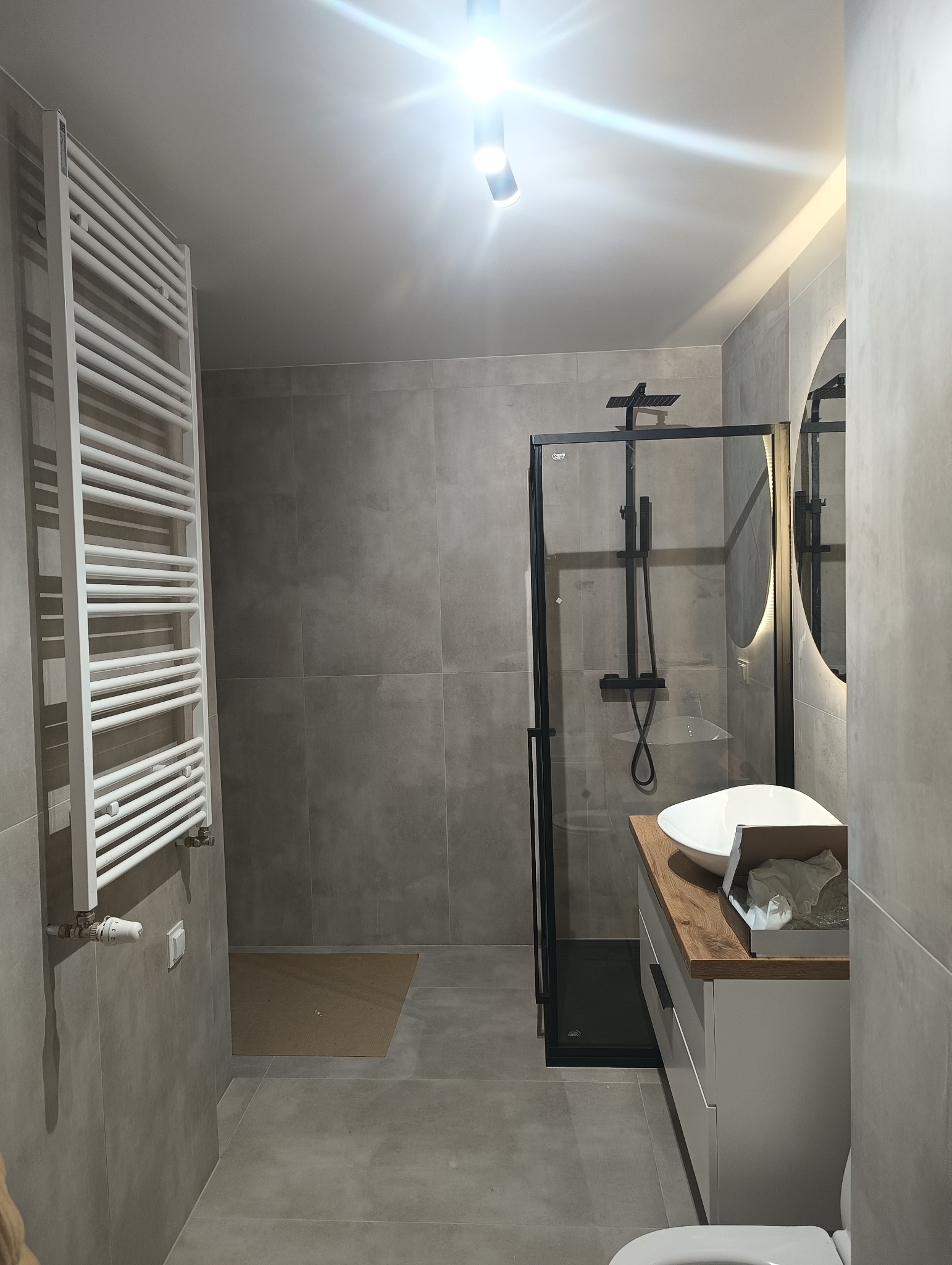 Nowoczesna łazienka z szarymi płytkami, kabiną prysznicową typu walk-in z czarnymi elementami, grzejnikiem drabinkowym, okrągłym lustrem z podświetleniem LED i umywalką nablatową na drewnianym blacie.