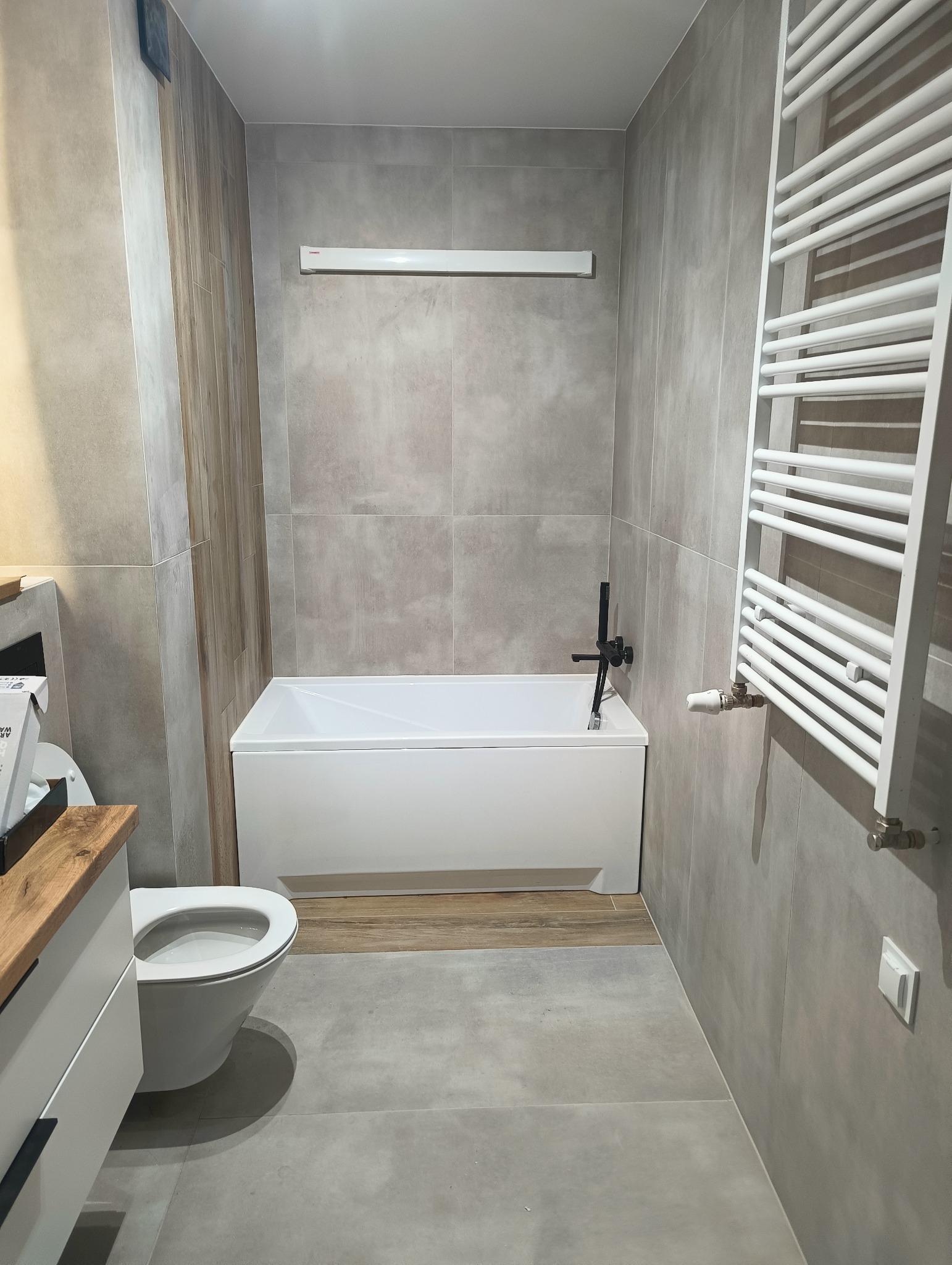 Nowoczesna łazienka po remoncie: szare płytki, biała wanna z czarną armaturą, grzejnik drabinkowy i podwieszana toaleta z drewnianym blatem.
