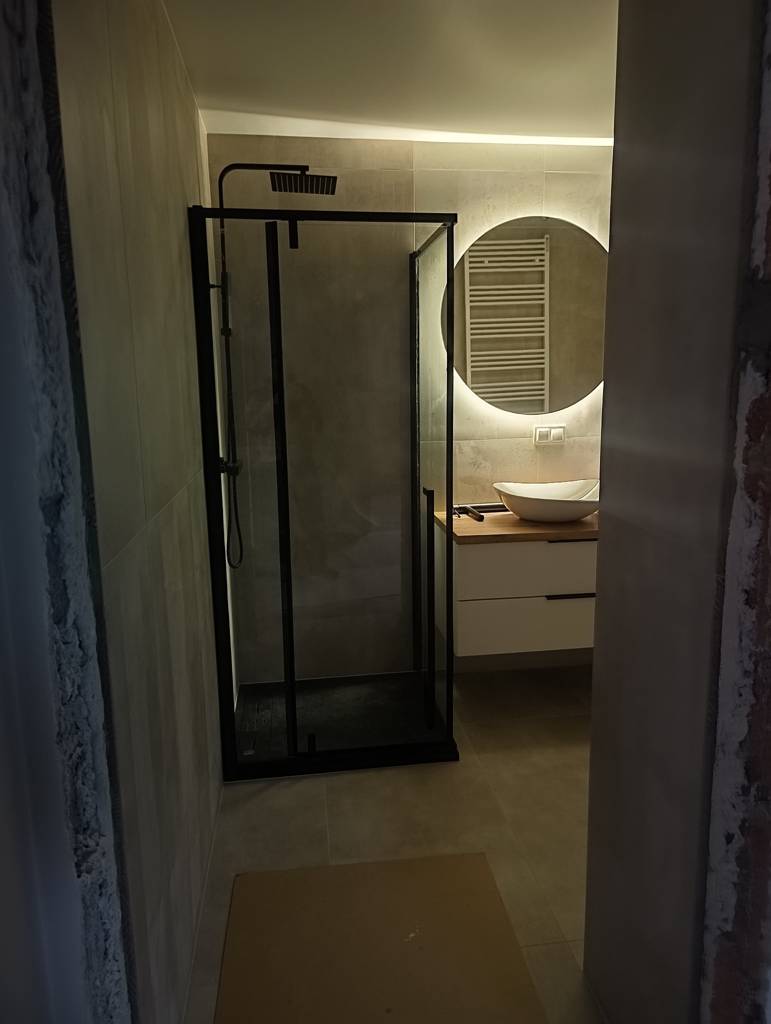Nowoczesna łazienka z czarną kabiną prysznicową typu walk-in, umywalką nablatową na drewnianym blacie i okrągłym lustrem z podświetleniem LED, widoczna z perspektywy wejścia do pomieszczenia.
