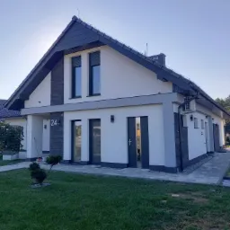 Nowoczesny dom jednorodzinny z białą elewacją, szarymi akcentami i numerem 24, otoczony zadbanym trawnikiem i ścieżką z kostki brukowej.