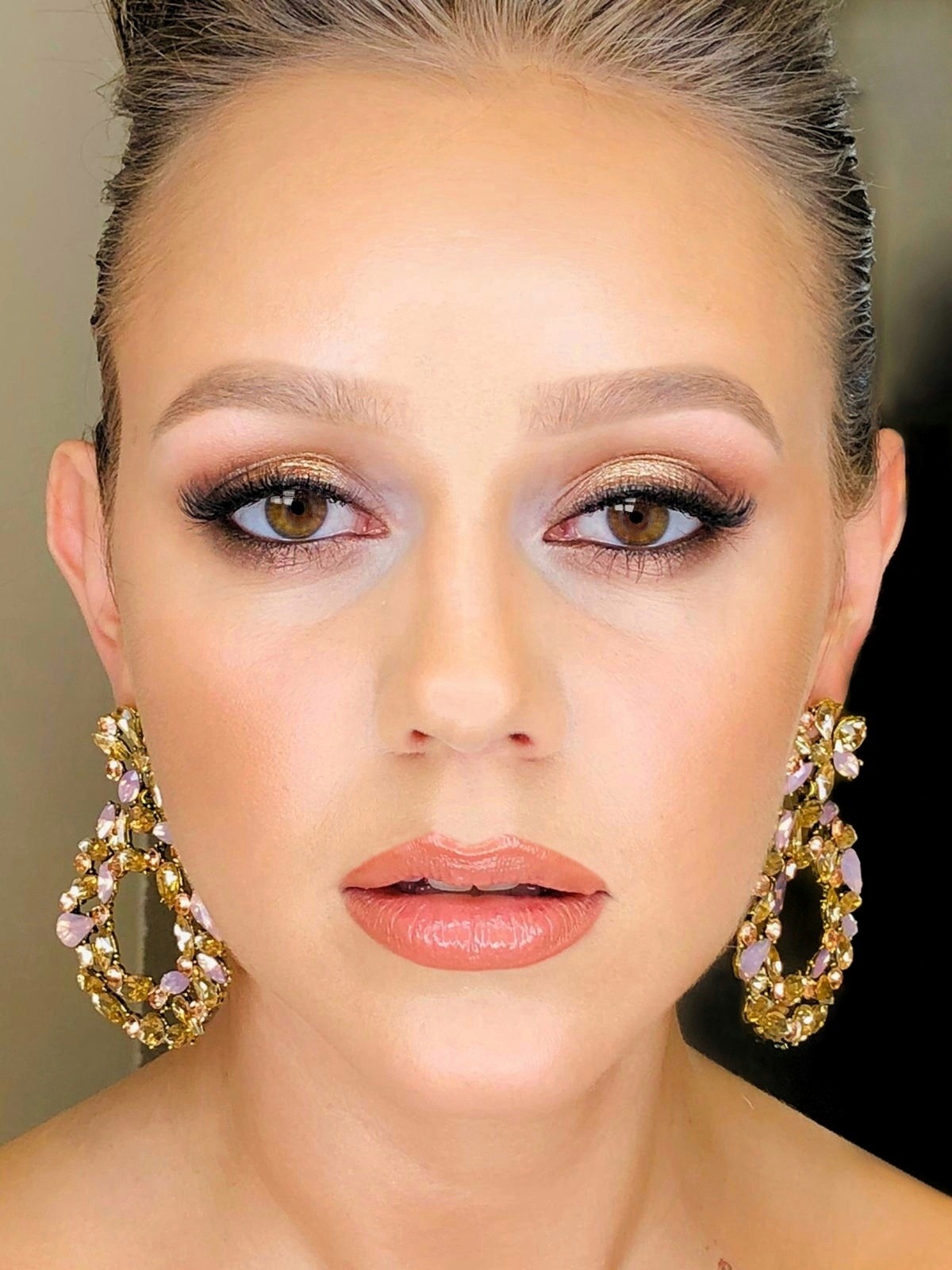 Portret kobiety z profesjonalnym makijażem oczu w odcieniach złota i brązu, podkreślonymi rzęsami, oraz błyszczącymi, pełnymi ustami. Uwagę przyciągają duże, zdobione kolczyki z kamieniami...