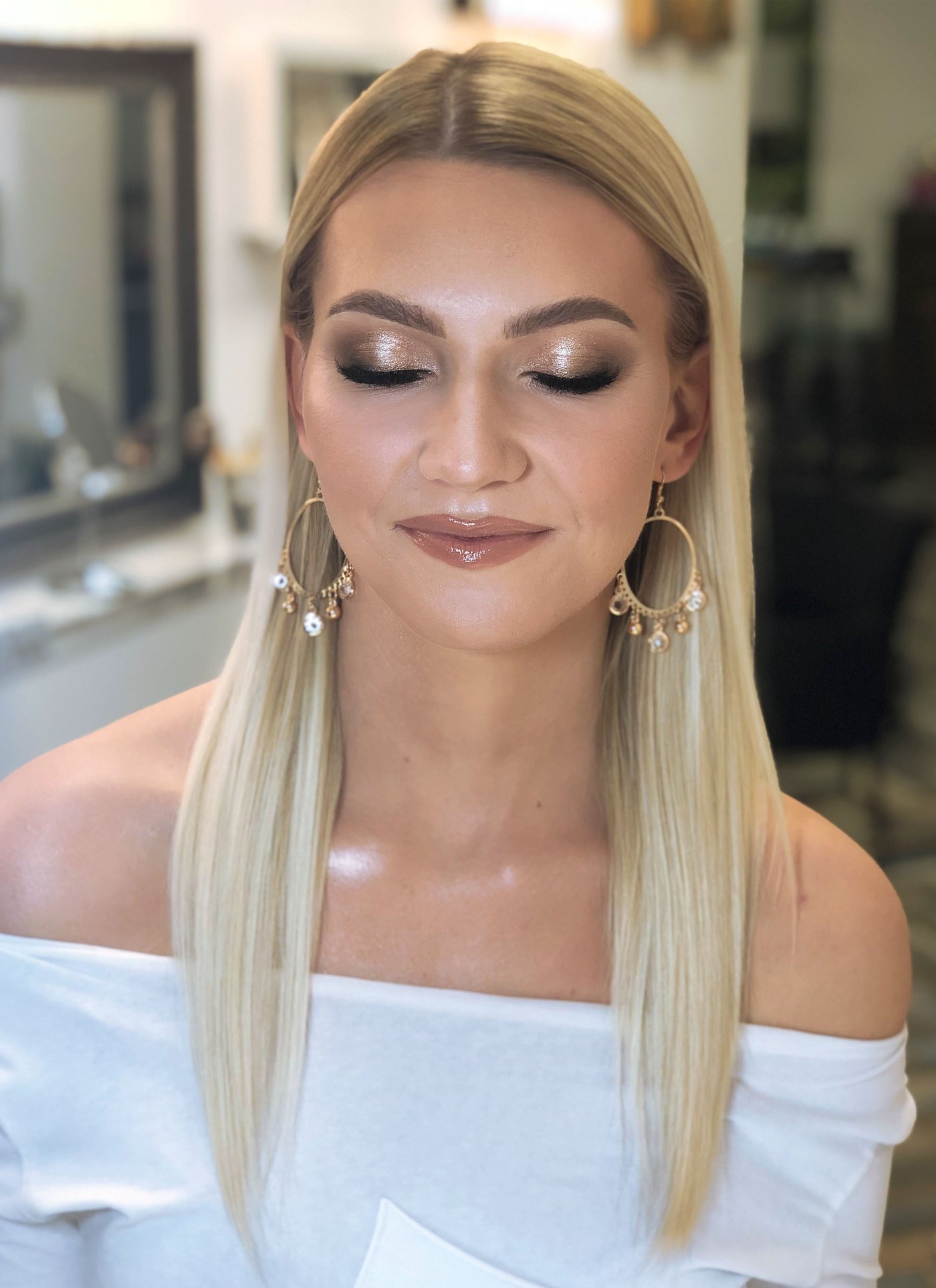 Portret kobiety z blond włosami i profesjonalnym makijażem oczu w odcieniach złota i brązu, błyszczące usta, duże kolczyki koła z wiszącymi ozdobami, zamknięte oczy.