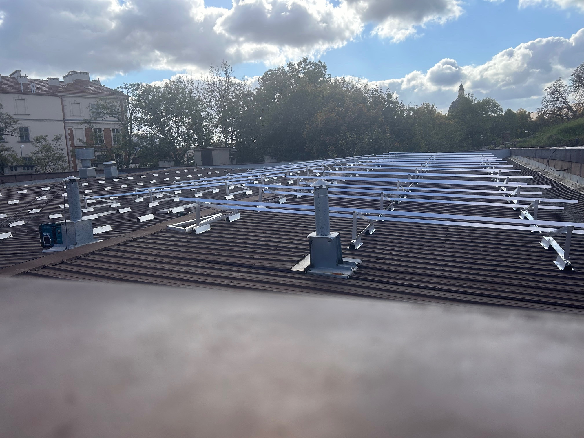 Montaż stelażu pod panele słoneczne na dachu z blachy trapezowej. Widoczne elementy mocujące i konstrukcja wsporcza. W tle budynki i drzewa pod błękitnym niebem.