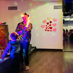 ANIMATOR Z KLASĄ PRZEMYSŁAW KORNIJENKO - Mężczyzna w koszuli w czaszki trzyma płonące pochodnie, mały chłopiec patrzy z bliska; w tle sala z dekoracjami i nagłośnieniem.