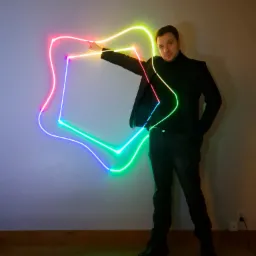 Mężczyzna w czarnym ubraniu prezentuje świetlny kształt przypominający nieregularny wielobok, emitujący światło w kolorach tęczy na tle szarej ściany.