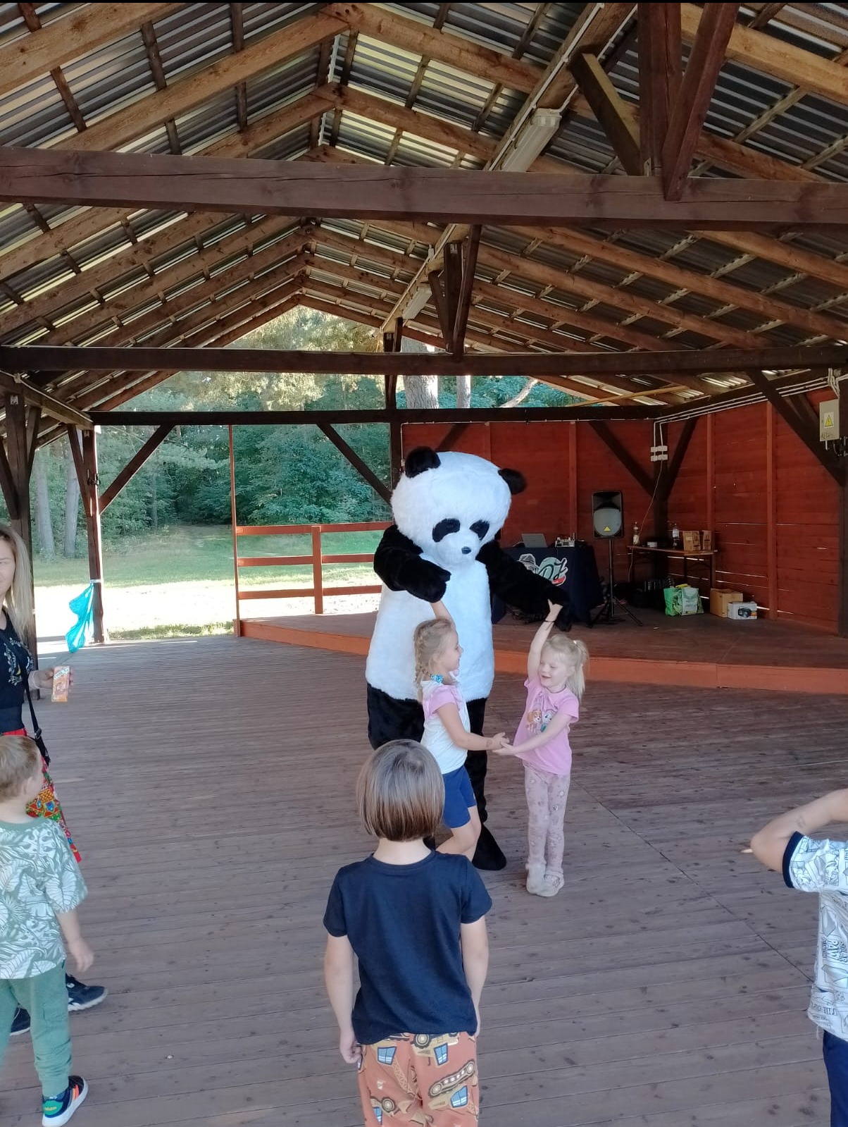 Osoba w kostiumie pandy tańczy z dwójką małych dziewczynek na drewnianej scenie pod zadaszeniem, w tle widoczny sprzęt nagłośnieniowy.