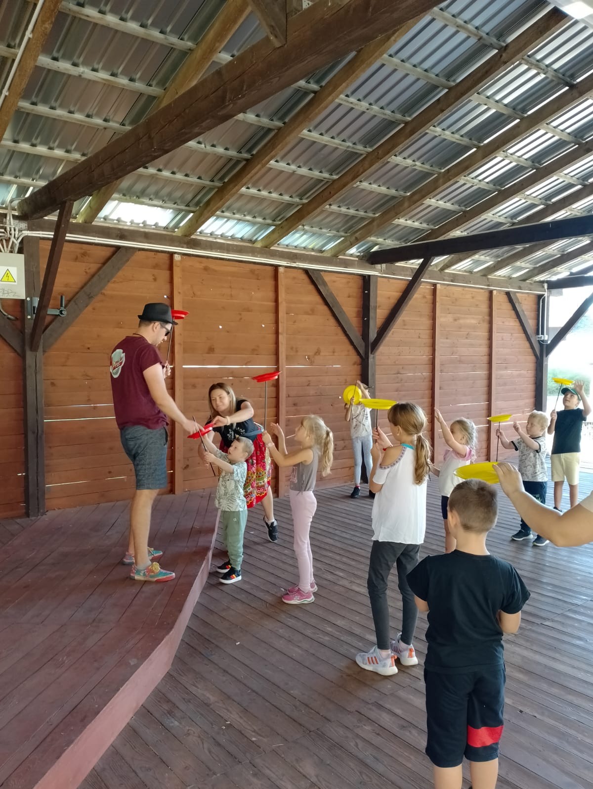 Instruktor w kapeluszu uczy dzieci kręcić talerzami na drewnianej scenie podczas festynu pod zadaszeniem.