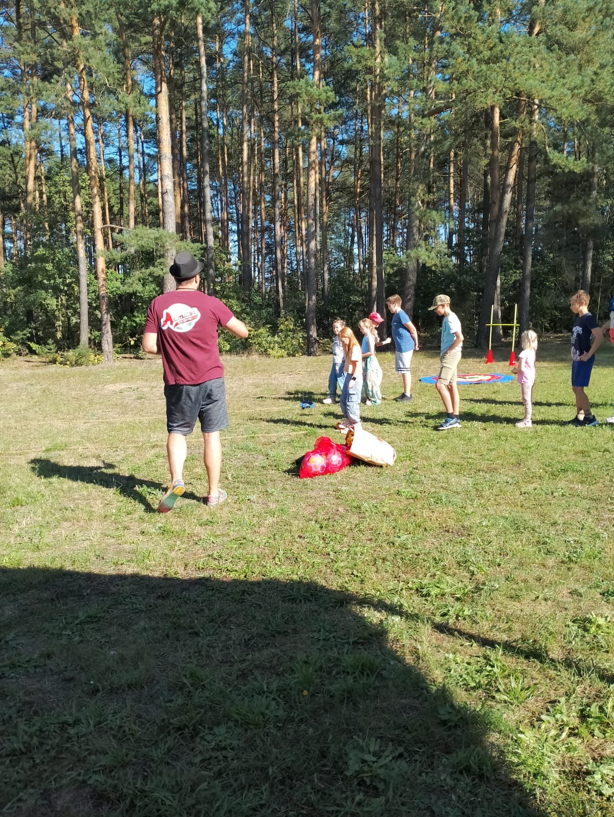 Grupa dzieci ustawiona w kolejce na trawie, obserwująca mężczyznę w kapeluszu i koszulce z nadrukiem, obok leżą czerwone balony i rekwizyty do zabawy, w tle wysoki las.