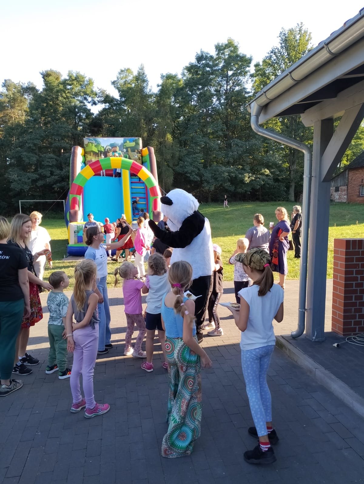 Dzieci otoczone przez dorosłych obserwują postać w kostiumie pandy na tle dmuchanego zamku ze zjeżdżalnią, podczas imprezy plenerowej.