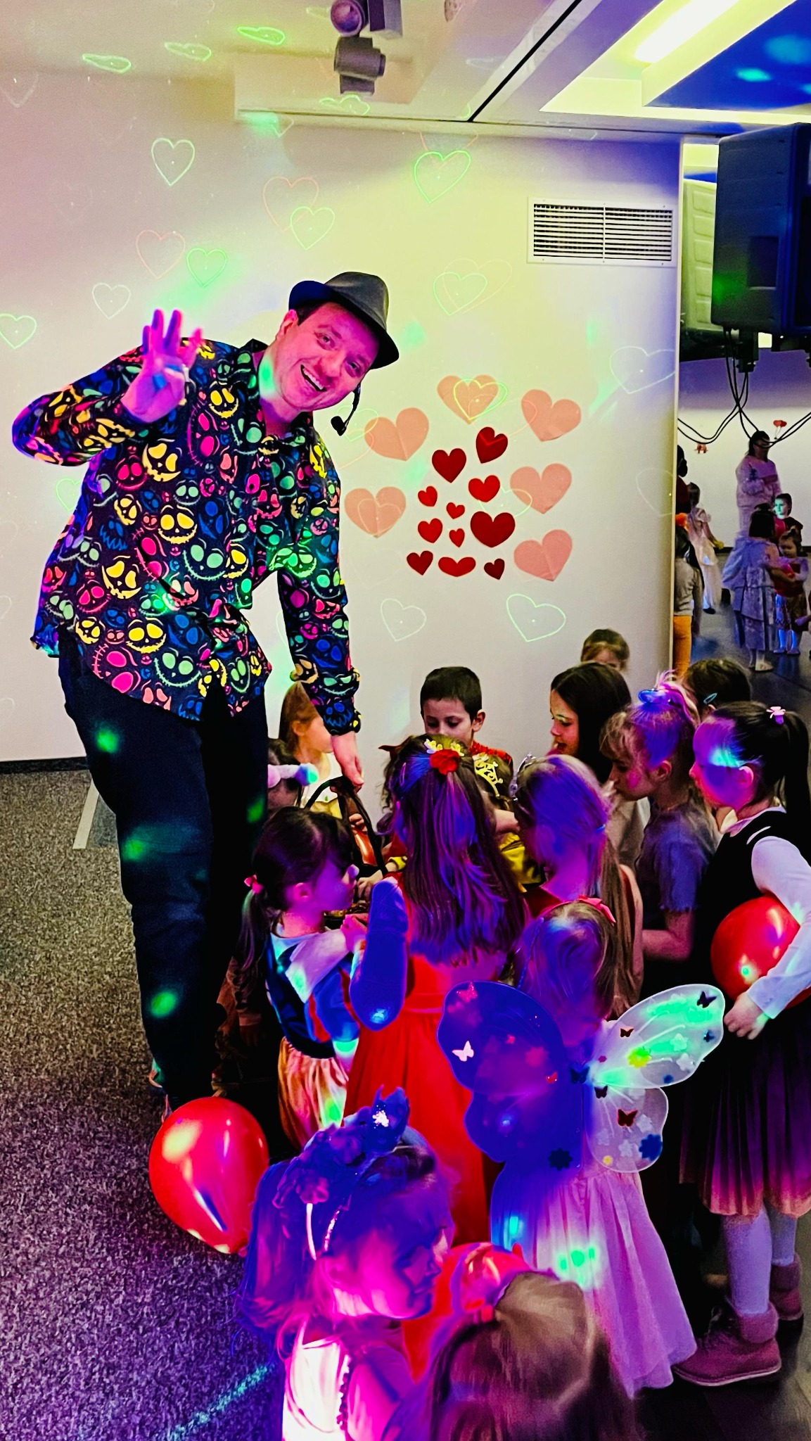 Uśmiechnięty mężczyzna w czarnym kapeluszu i koszuli w kolorowe czaszki, z mikrofonem nagłownym, macha ręką do grupy dzieci w kostiumach, oświetlonych kolorowymi światłami i projekcjami serc...