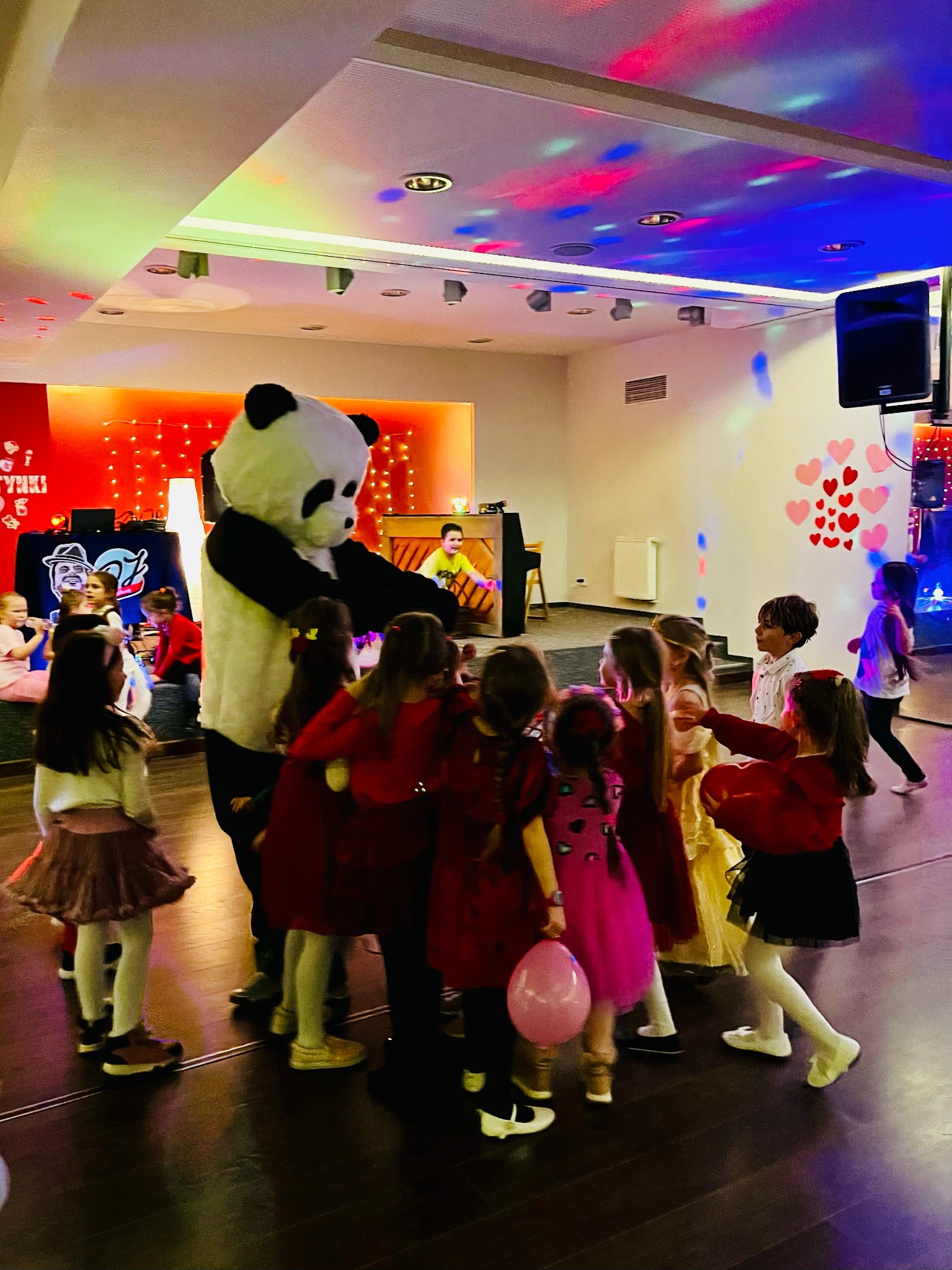Panda w kostiumie otoczona grupą dzieci w sali z dekoracjami, w tle pianino i osoba grająca.