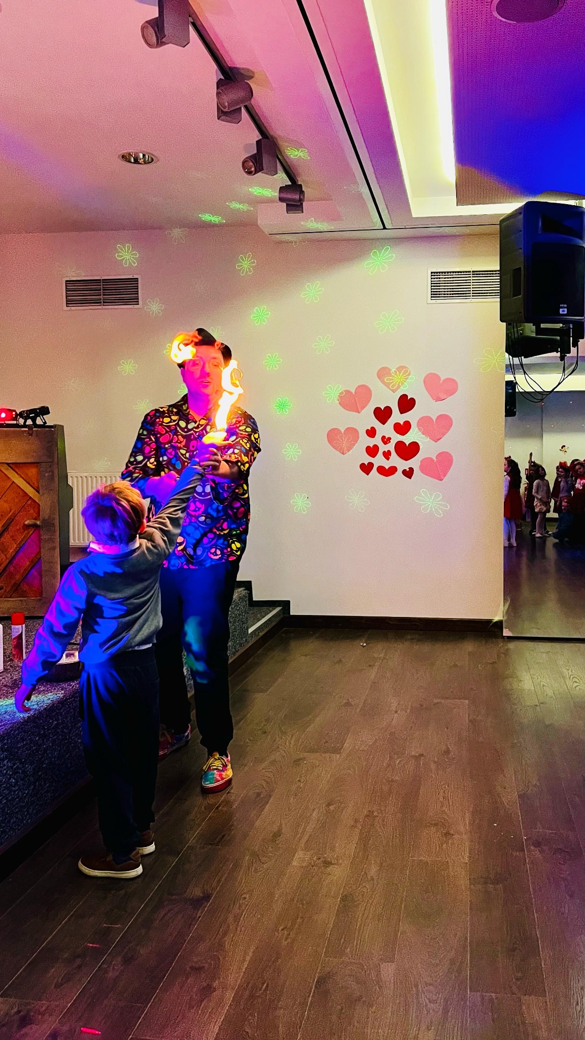 Mężczyzna w koszuli w czaszki trzyma płonące pochodnie, mały chłopiec patrzy z bliska; w tle sala z dekoracjami i nagłośnieniem.