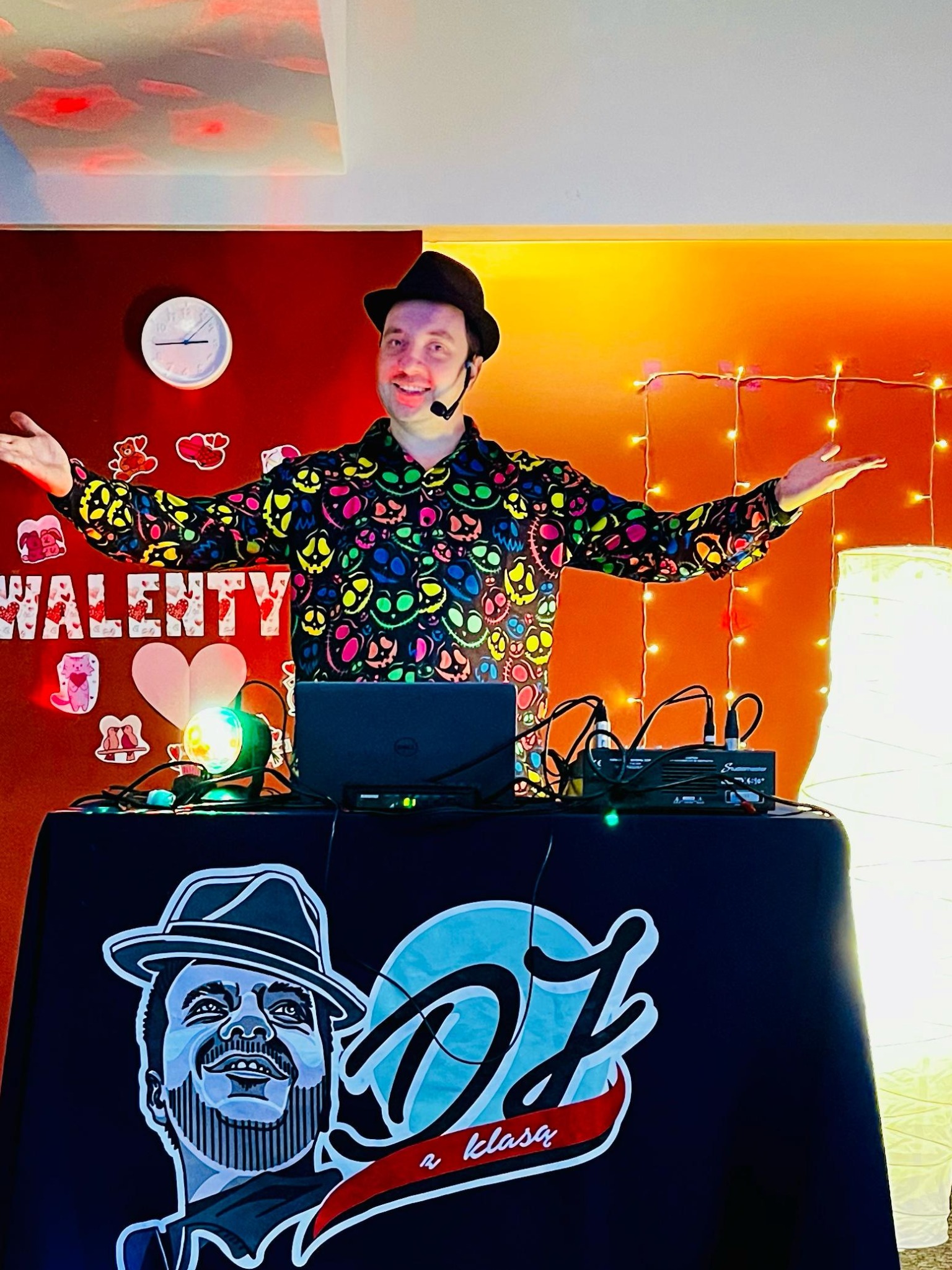 Uśmiechnięty DJ/Animator w czarnym kapeluszu i koszuli w kolorowe czaszki, z mikrofonem nagłownym, rozkłada ręce na tle dekoracji walentynkowych i sprzętu DJ-skiego.