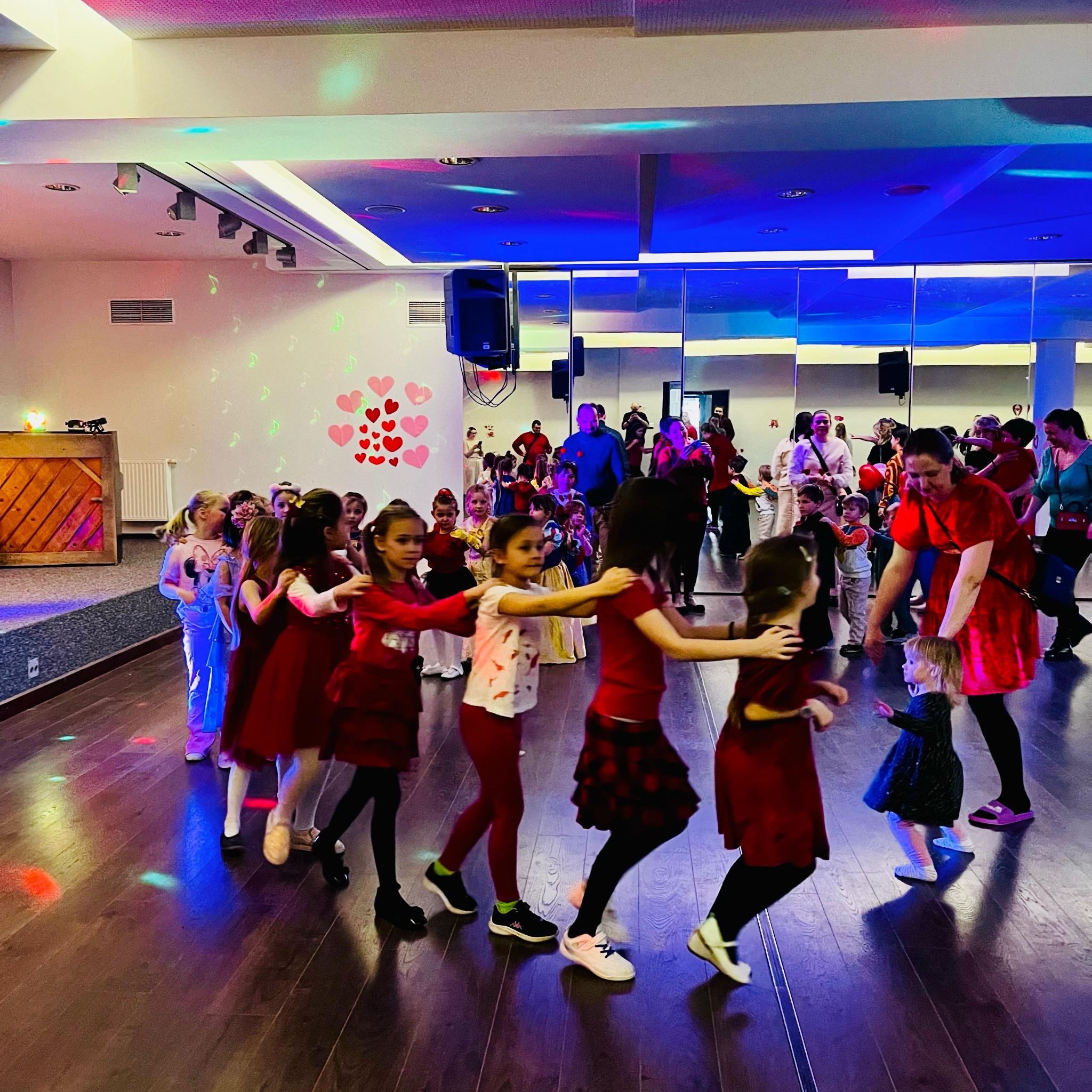 Grupa dzieci w różnym wieku, ubranych w stroje, tańczy w rzędzie na parkiecie oświetlonym kolorowymi światłami, pod nadzorem dorosłej kobiety w czerwonej sukience. W tle widoczna scena, lustra...