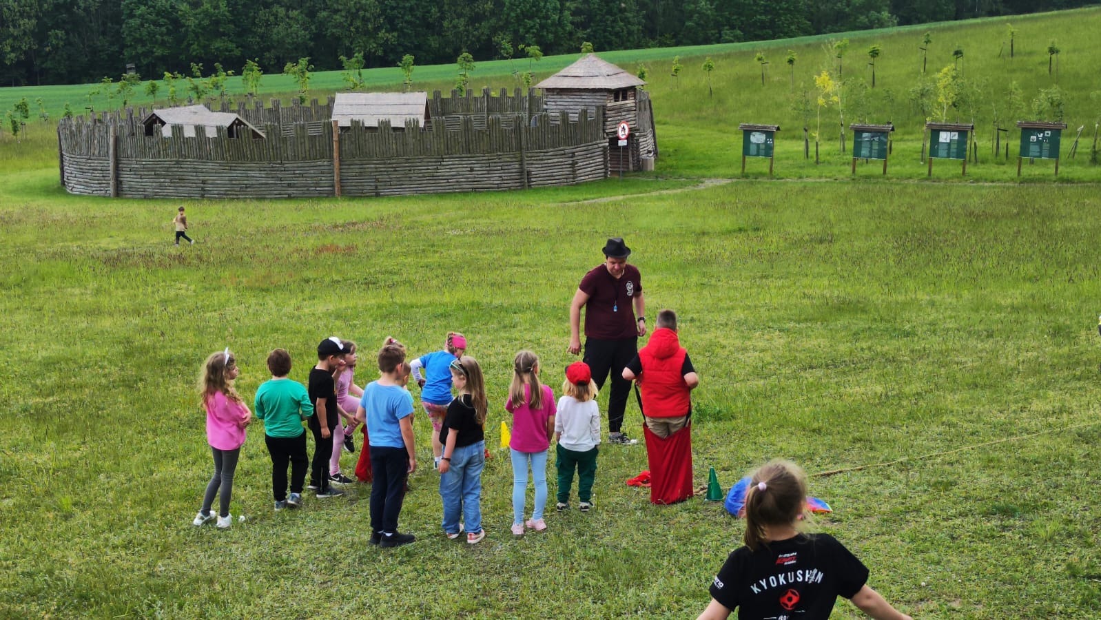 Animator prowadzi gry i zabawy dla grupy dzieci na zielonej łące z drewnianą konstrukcją w tle, w plenerze. Dzieci w różnym wieku i strojach, animator w kapeluszu.