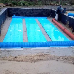 Instalacja rekuperacji gruntowej: niebieskie rury karbowane i płaskie kolektory ułożone na piasku w wykopie fundamentowym, widoczny pracownik przy laptopie.