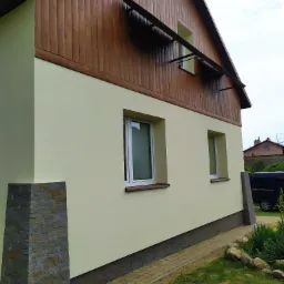 Elewacja domu jednorodzinnego z jasnym tynkiem i drewnianą boazerią na poddaszu, z kamienną okładziną narożnika i widoczną rynną na górze oraz kostką brukową wokół budynku.