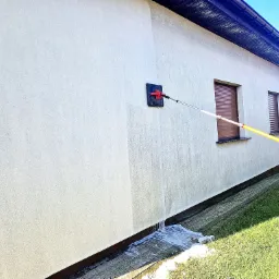 Elewacja budynku jednorodzinnego czyszczona myjką z długim, żółtym teleskopowym uchwytem, woda spływa po ścianie na chodnik.
