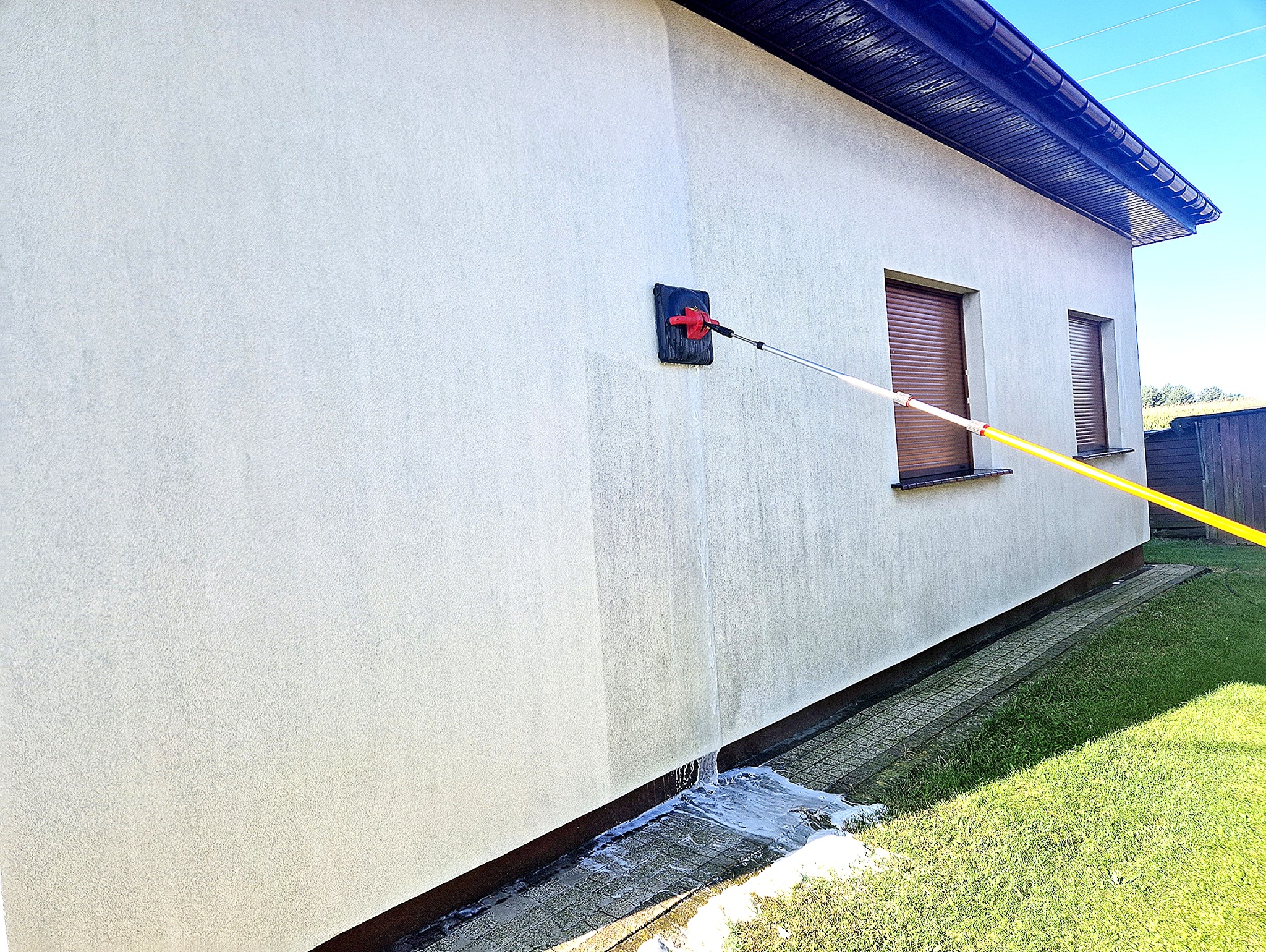 Elewacja budynku jednorodzinnego czyszczona myjką z długim, żółtym teleskopowym uchwytem, woda spływa po ścianie na chodnik.