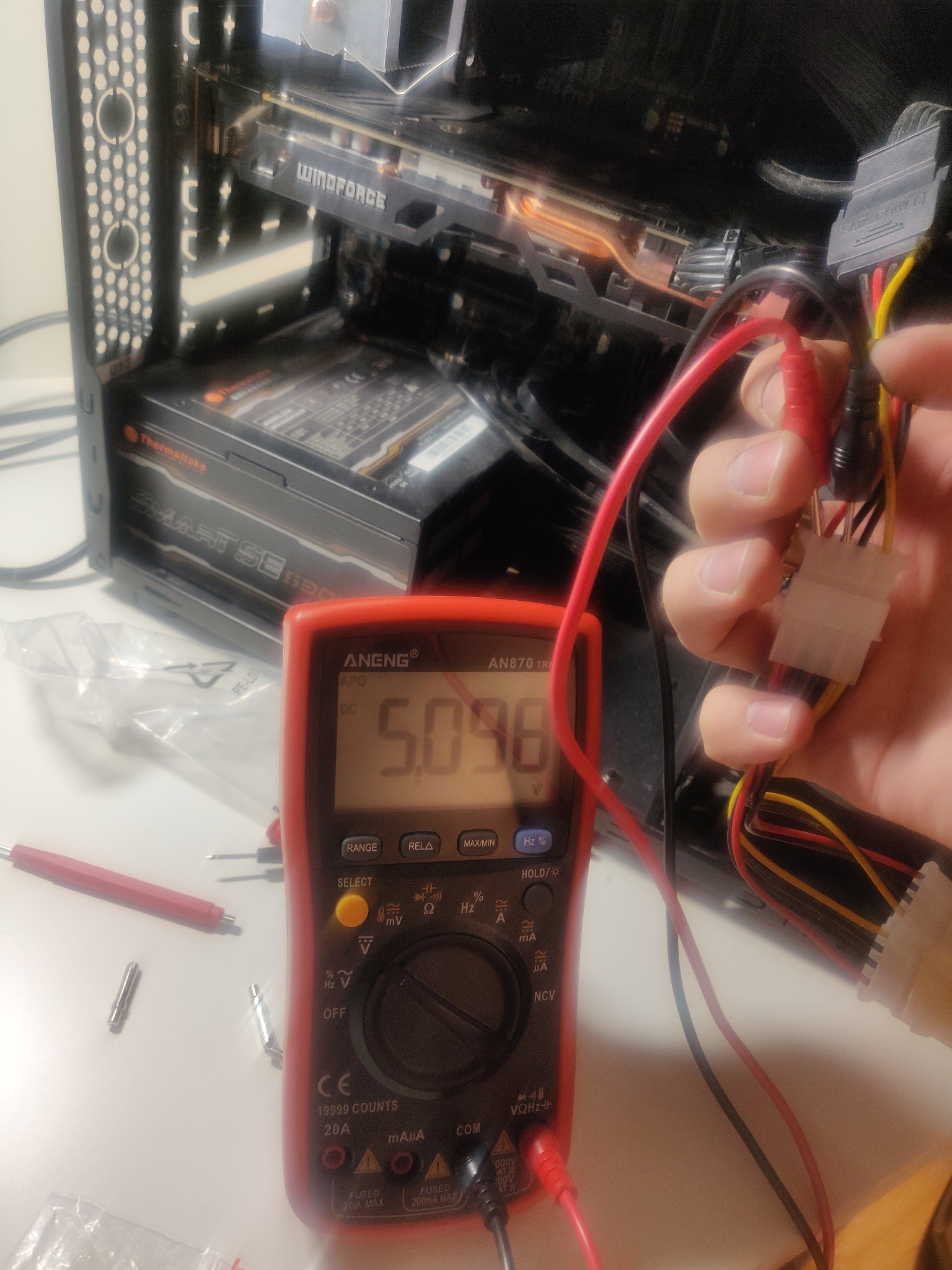 Sprawdzanie napięcia zasilacza komputerowego multimetrem ANENG AN870 TR podczas naprawy komputera, widoczny otwarty komputer z kartą graficzną Windforce i zasilacz Thermaltake Smart SE 530.