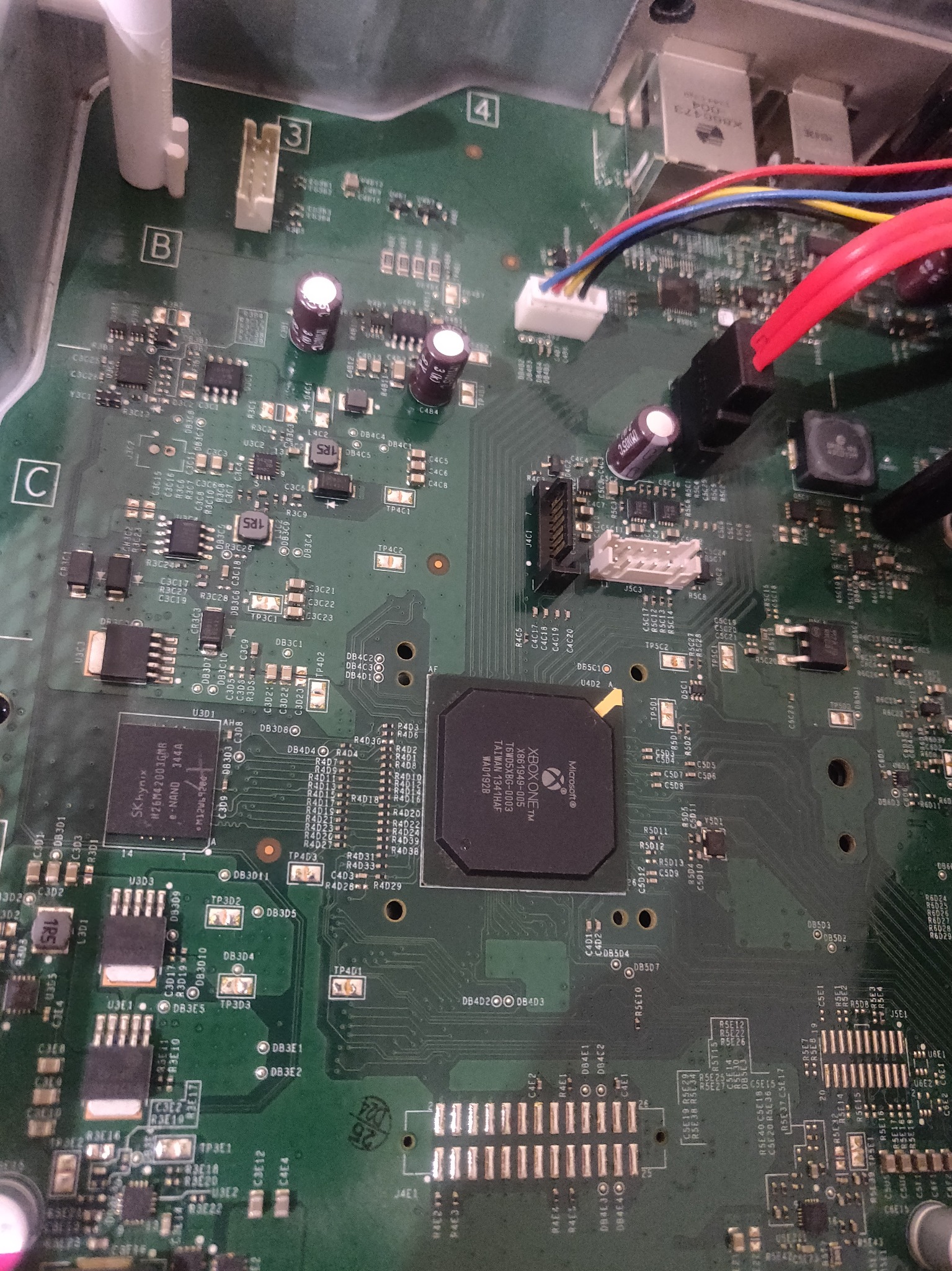 Widok z bliska płyty głównej konsoli Xbox One z widocznymi elementami elektronicznymi, takimi jak chipy, kondensatory i złącza, z przewodami podłączonymi do jednego ze złącz.