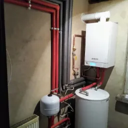 Biały piec grzewczy Viessmann z podłączonymi rurami miedzianymi i czerwonymi, szarymi rurami izolacyjnymi, białym zasobnikiem ciepłej wody i grzejnikiem żeberkowym na tle beżowej ściany.