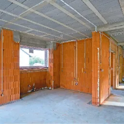 Surowy stan wnętrza domu w trakcie budowy: widoczne ściany z czerwonej cegły, betonowy sufit z widocznymi belkami, częściowo rozprowadzone przewody elektryczne i instalacje hydrauliczne, okno...