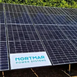 Rząd paneli słonecznych z logo Nortmar Power Systems, zainstalowanych na ziemi, z zielonymi krzewami w tle.