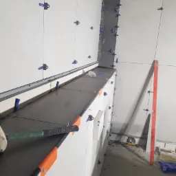 Montaż płytek wielkoformatowych na ścianie, użycie systemu poziomowania z niebieskimi klipsami, widoczna poziomica i gumowy młotek.