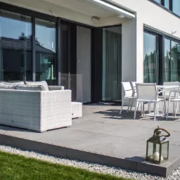 Nowoczesny taras z dużymi przesuwnymi oknami, białymi meblami ogrodowymi i dekoracyjną latarnią, widok na zadbany trawnik.