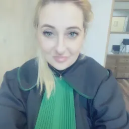 Portret kobiety w todze adwokackiej z zielonym żabotem, delikatny uśmiech, w tle jasne biuro z drewnianymi meblami.