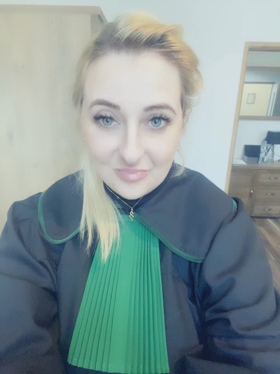 Portret kobiety w todze adwokackiej z zielonym żabotem, delikatny uśmiech, w tle jasne biuro z drewnianymi meblami.