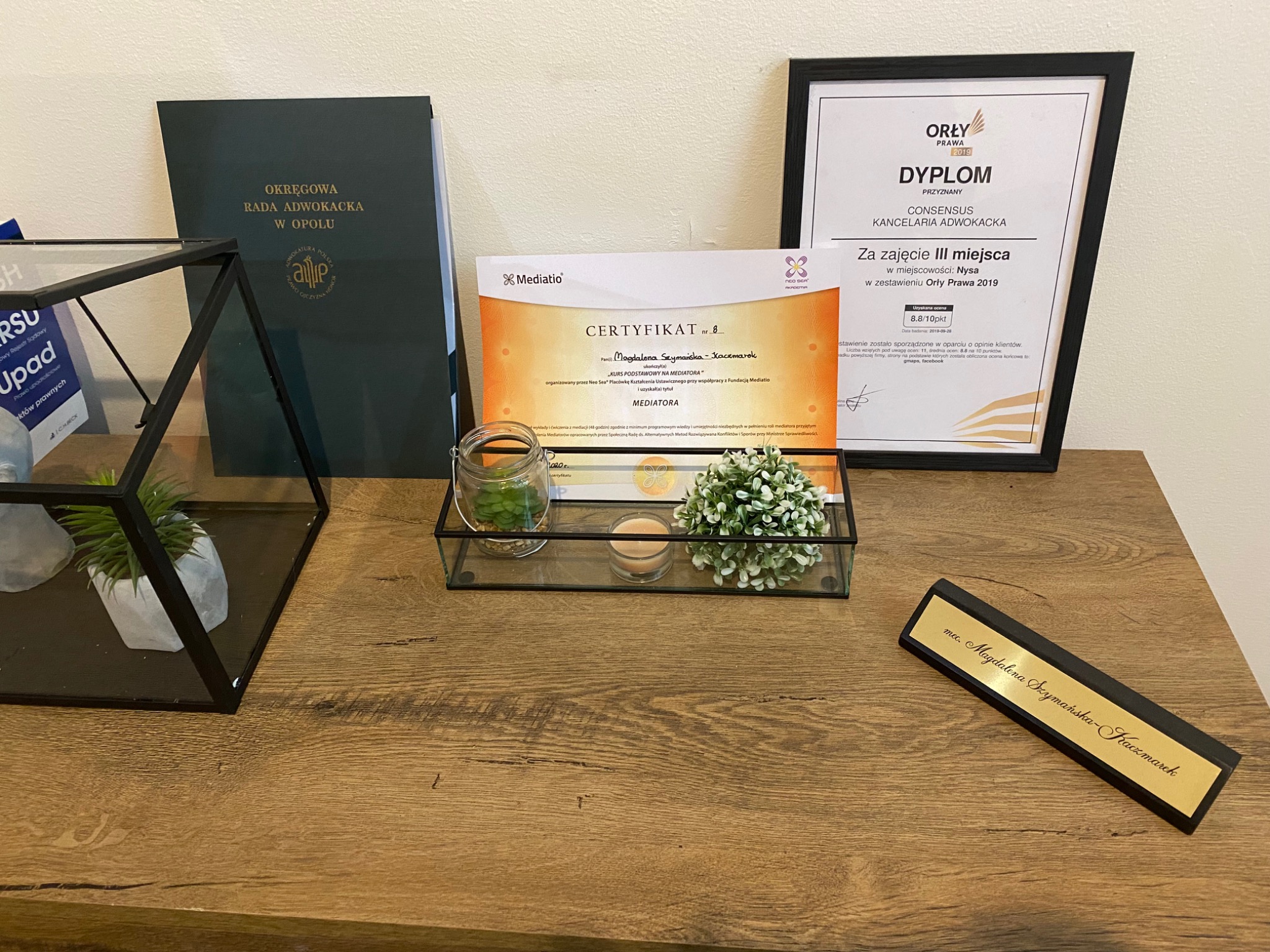 Gabinet adwokacki: dyplom za zajęcie III miejsca Orły Prawa 2019, certyfikat mediatora, tabliczka z imieniem i nazwiskiem adwokata, roślina w szklanej doniczce i dekoracje na drewnianym biurku.