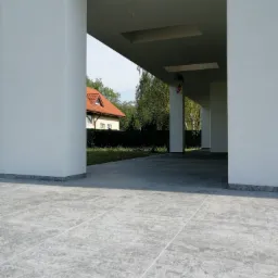 Zadaszony taras z szarych płytek ceramicznych, widok na ogród z domem w tle.